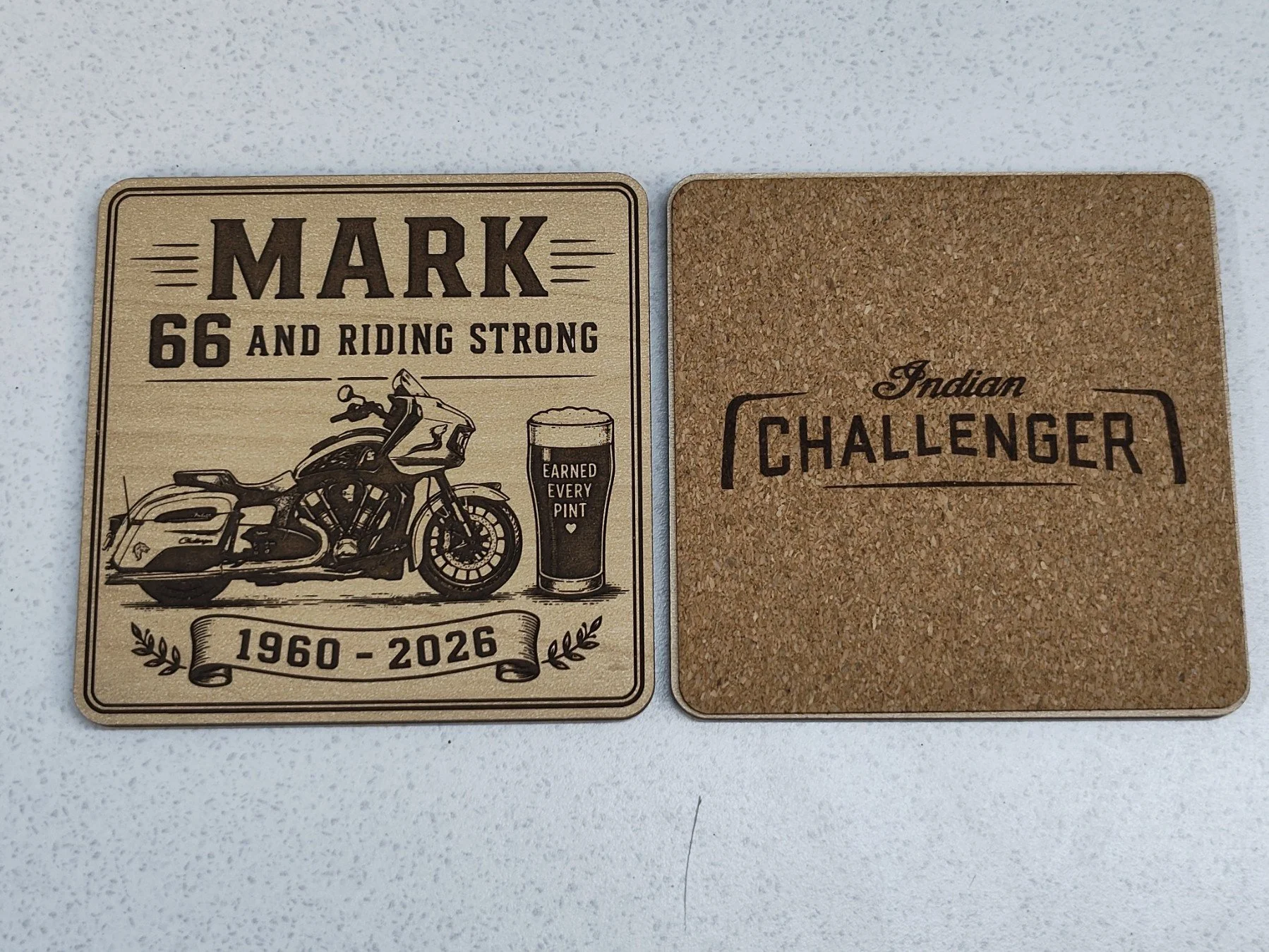 Two square coasters, one with a vintage motorcycle and a pint glass and the words "Mark 66 and Riding Strong," along with the years 1960-2026, and the other with the words "Indian Challenger" on a cork background.