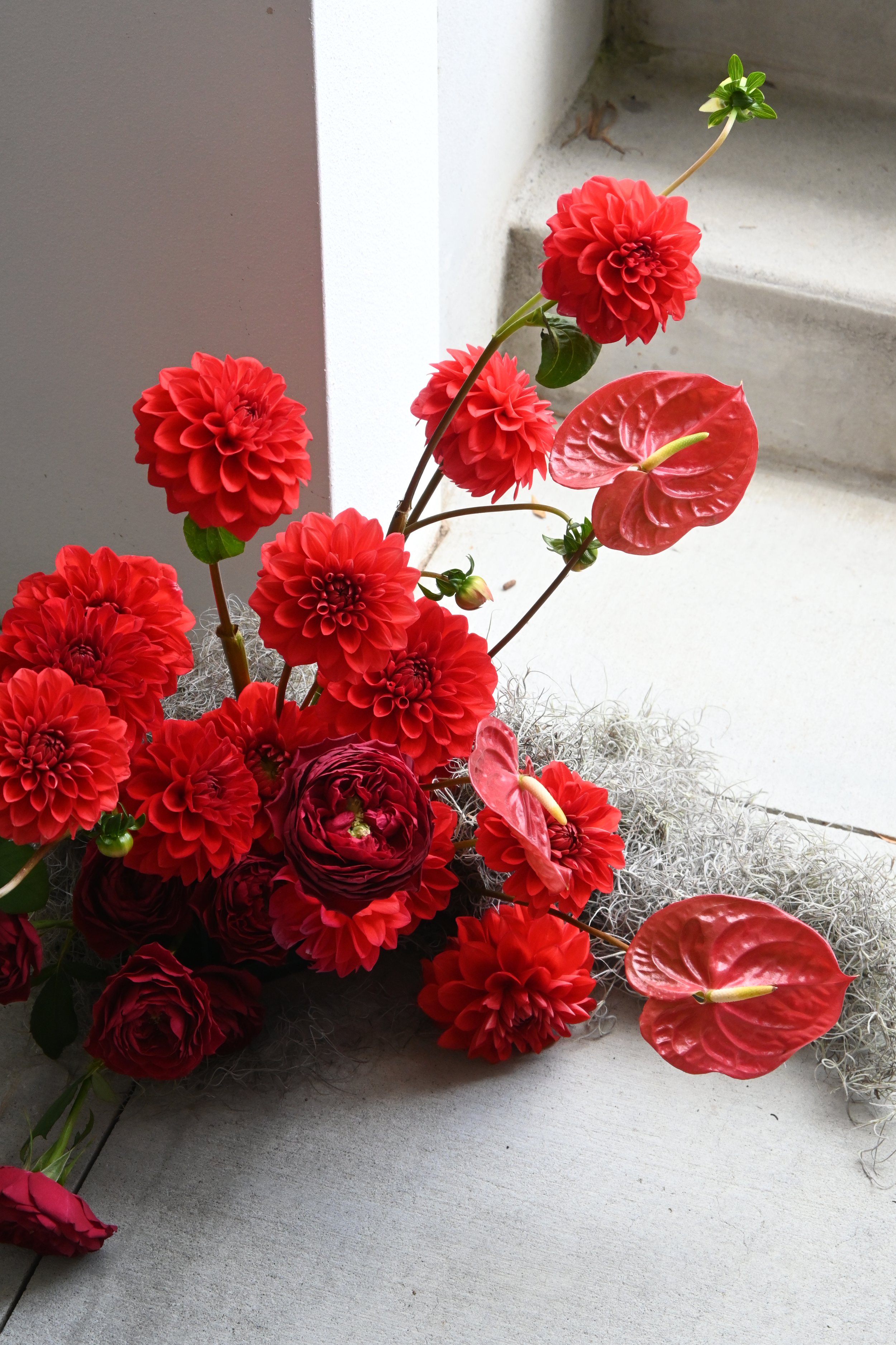 A floral arrangement with red dahlias, red anthuriums, and a few green leaves on a white surface near a window.