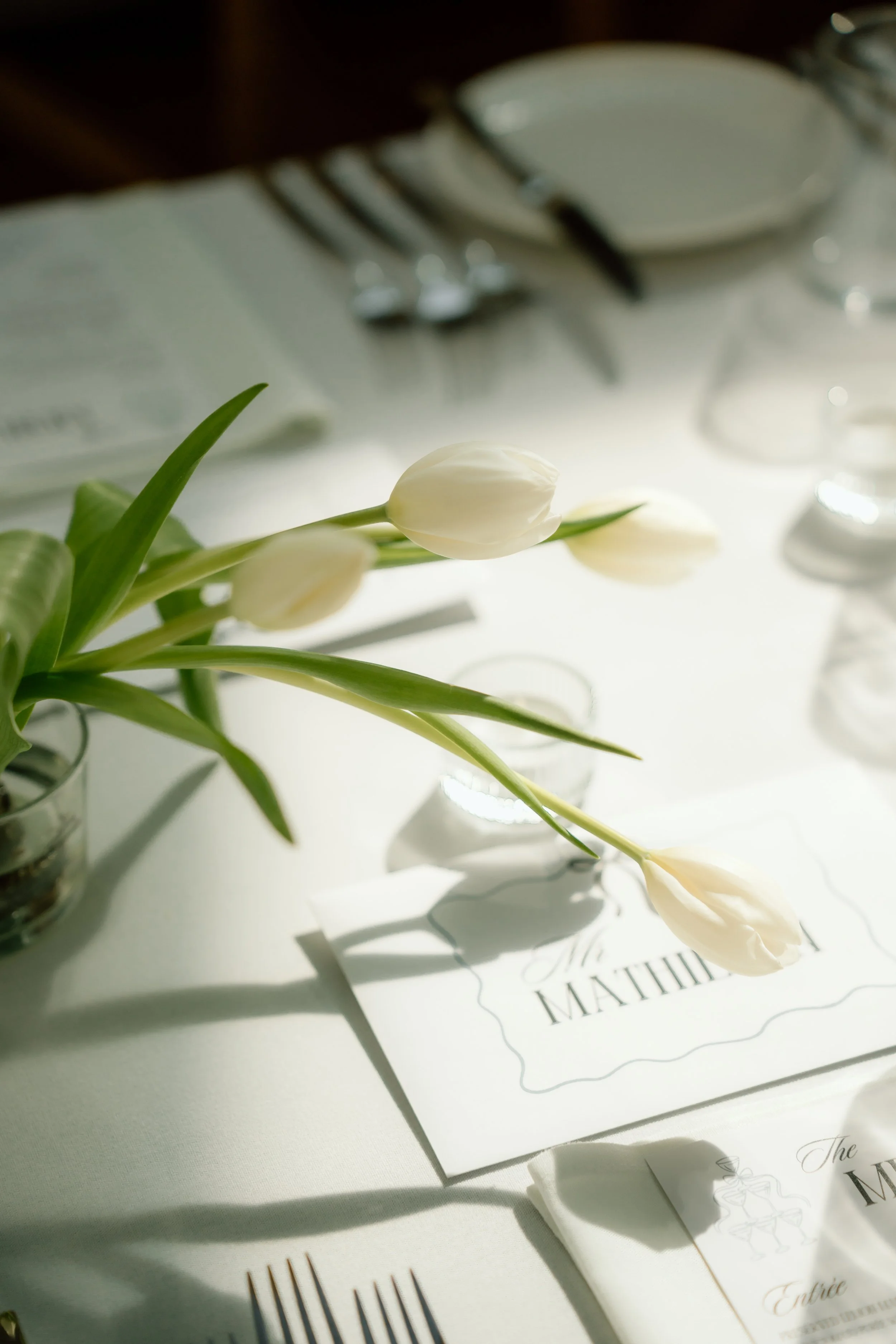 White tulips on a table with a mat or place card, silverware, plates, and glassware, set for a formal event or wedding reception.