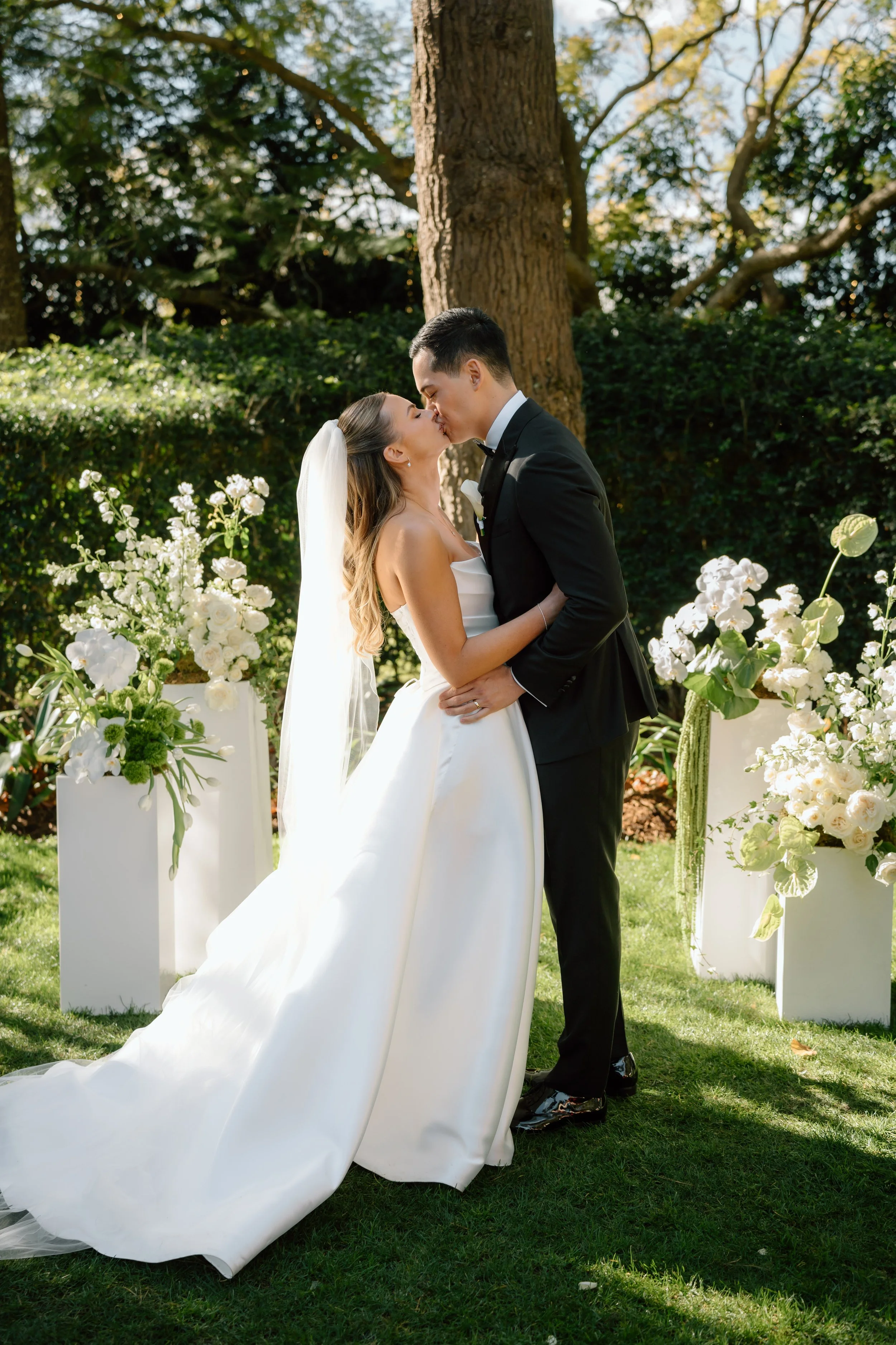 A bride and groom kissing outdoors during their wedding ceremony, with floral arrangements and a large tree in the background.