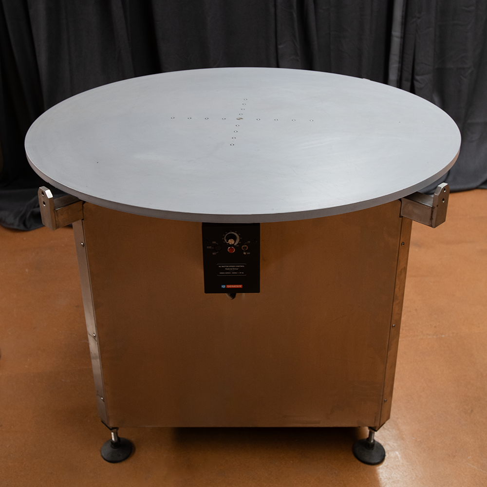A large circular electric heating or cooking appliance with a grey top and a control panel on the front. The appliance has a stainless steel base and is set on wheels or adjustable feet. The background is dark curtains.