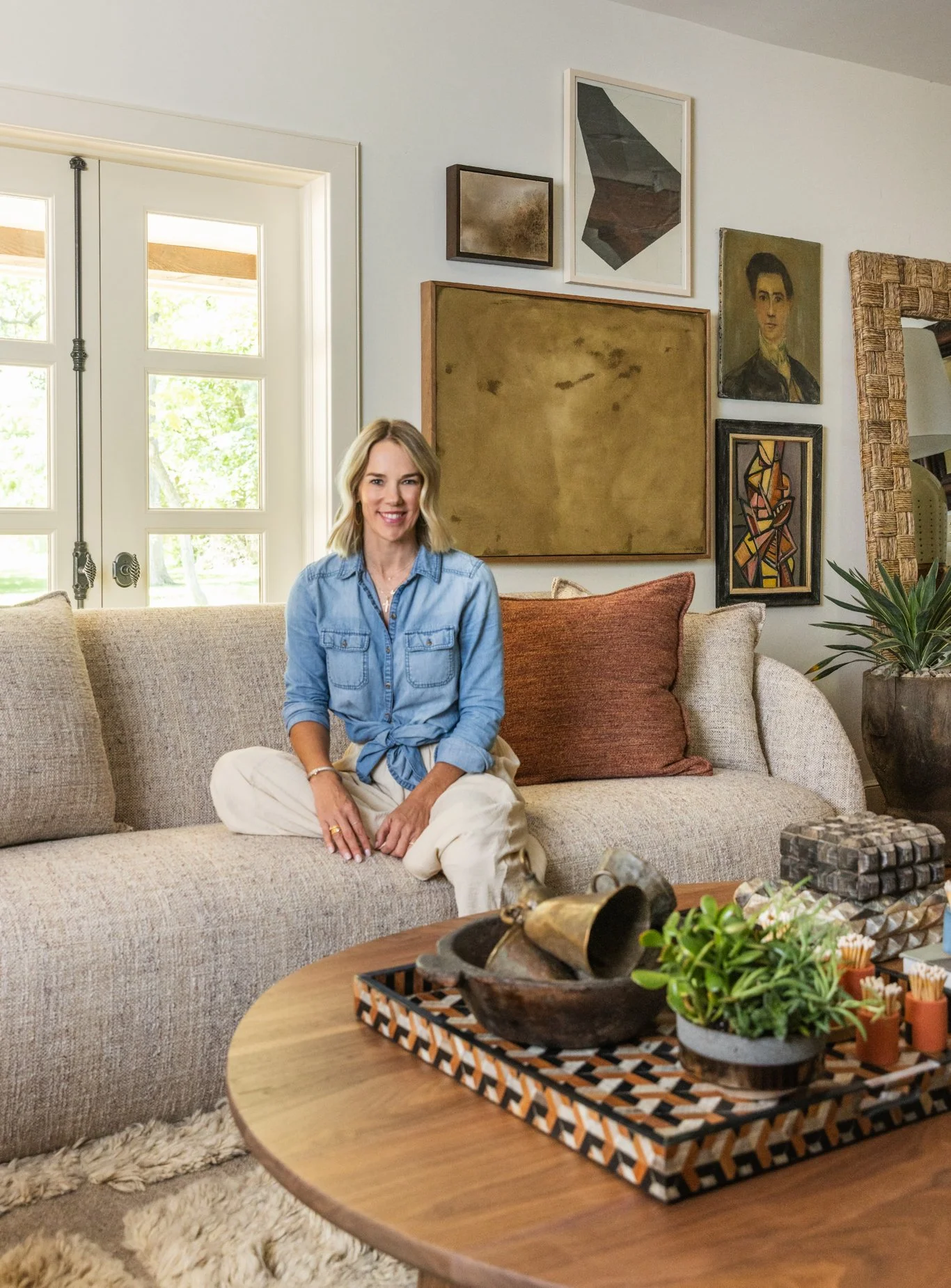 A woman sitting on a beige sofa in a cozy, art-filled living room, smiling at the camera.