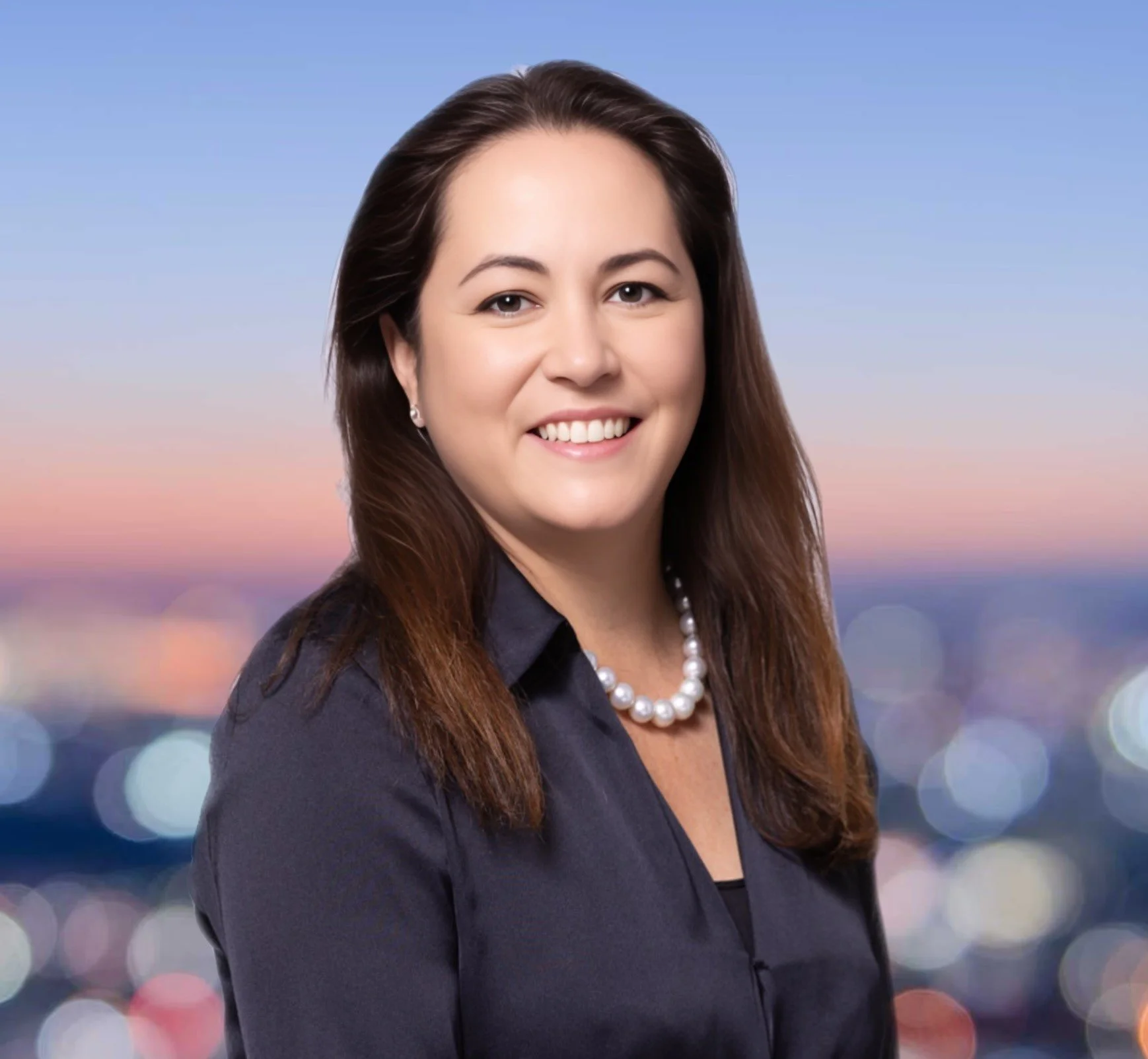 Portrait of a smiling woman with brown hair, wearing a dark blazer and pearl necklace, against a blurred cityscape background at sunset.