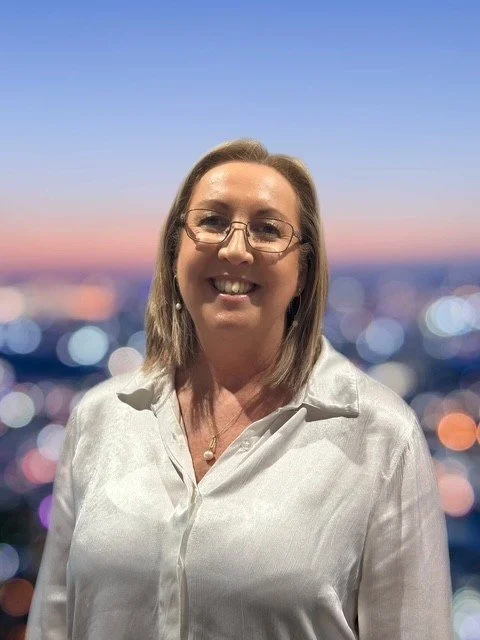 A middle-aged woman with glasses and shoulder-length hair smiling in front of a blurry cityscape at sunset, wearing a white blouse.