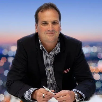 A man sitting at a desk, holding a pen, with a blurred cityscape background during sunset.