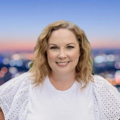 A woman with wavy blonde hair smiling, wearing a white top with eyelet sleeves, standing in front of a blurred cityscape at sunset.