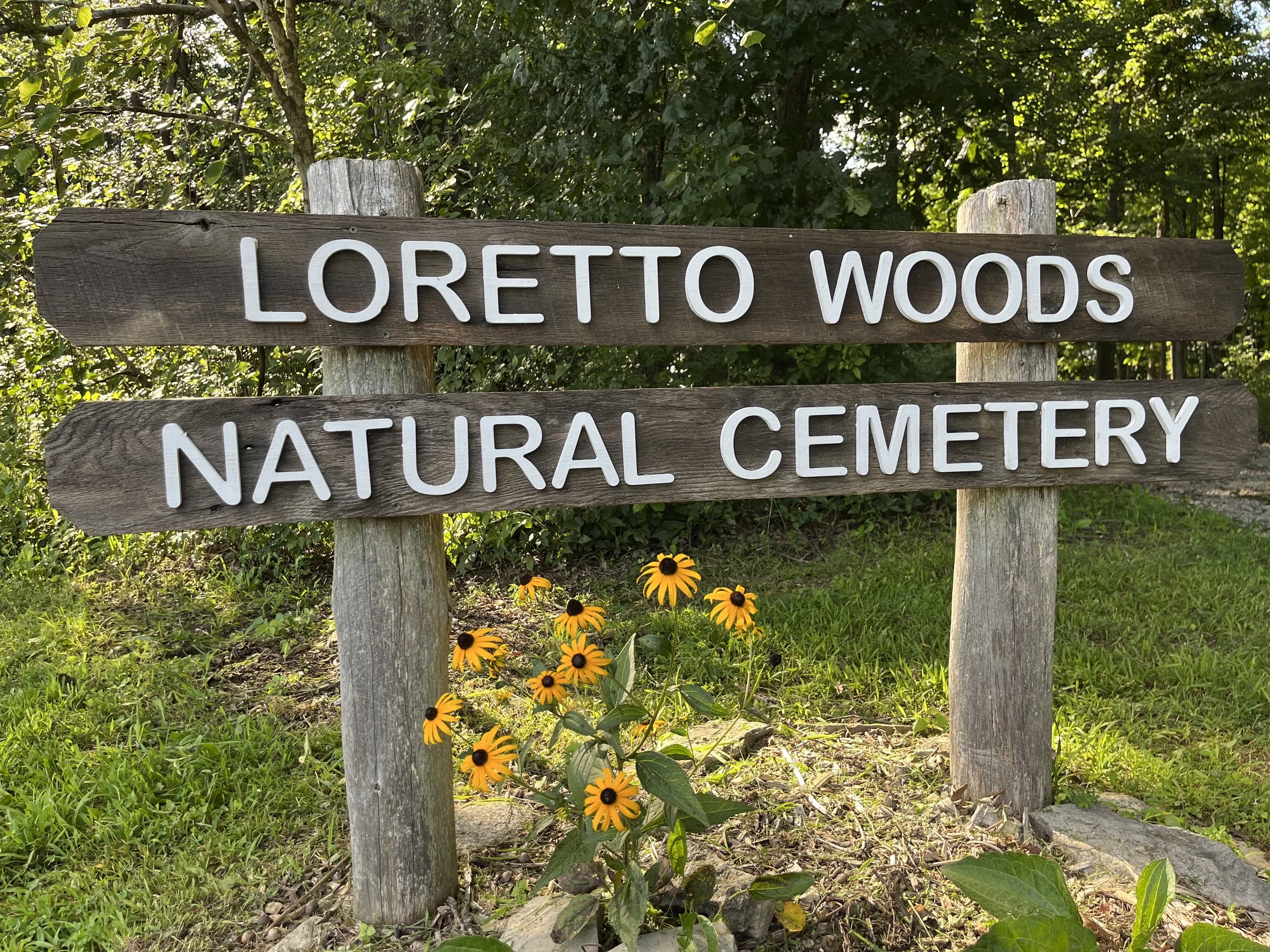 A wooden sign that reads "Loretto Woods Natural Cemetery" in front of lush, green trees with yellow rudbeckia flowers growing beneath.