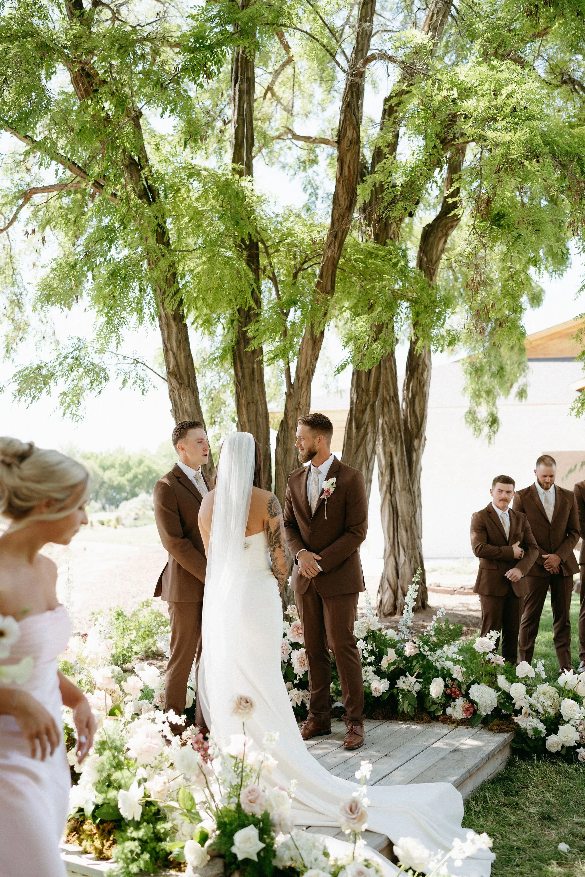 A wedding ceremony outdoors under large green trees, with a bride and groom standing at the altar surrounded by floral arrangements, and bridesmaids and groomsmen dressed in brown suits.