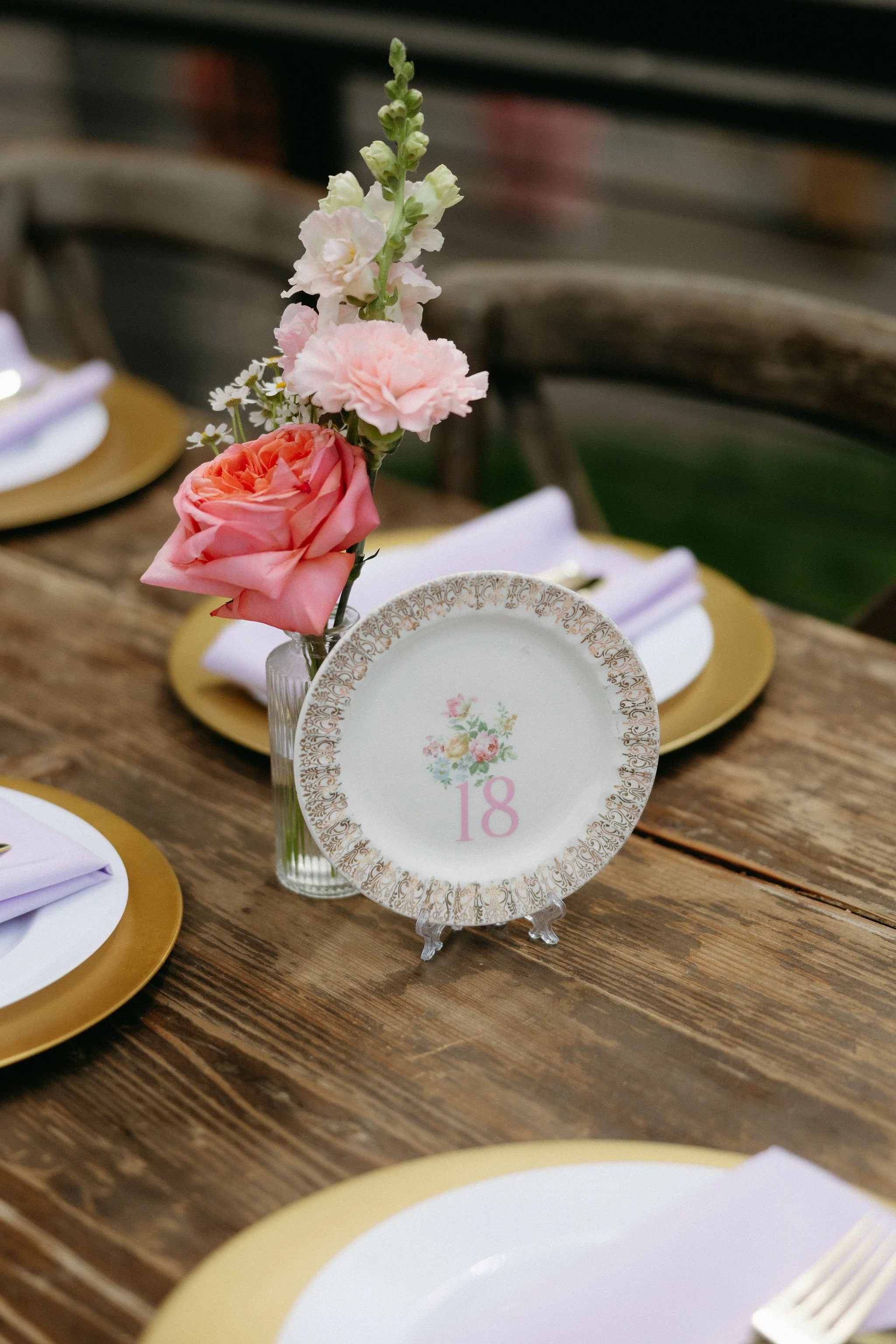 A floral centerpiece with pink and white flowers in a small glass vase on a wooden table, with a decorative table card displaying the number 18.