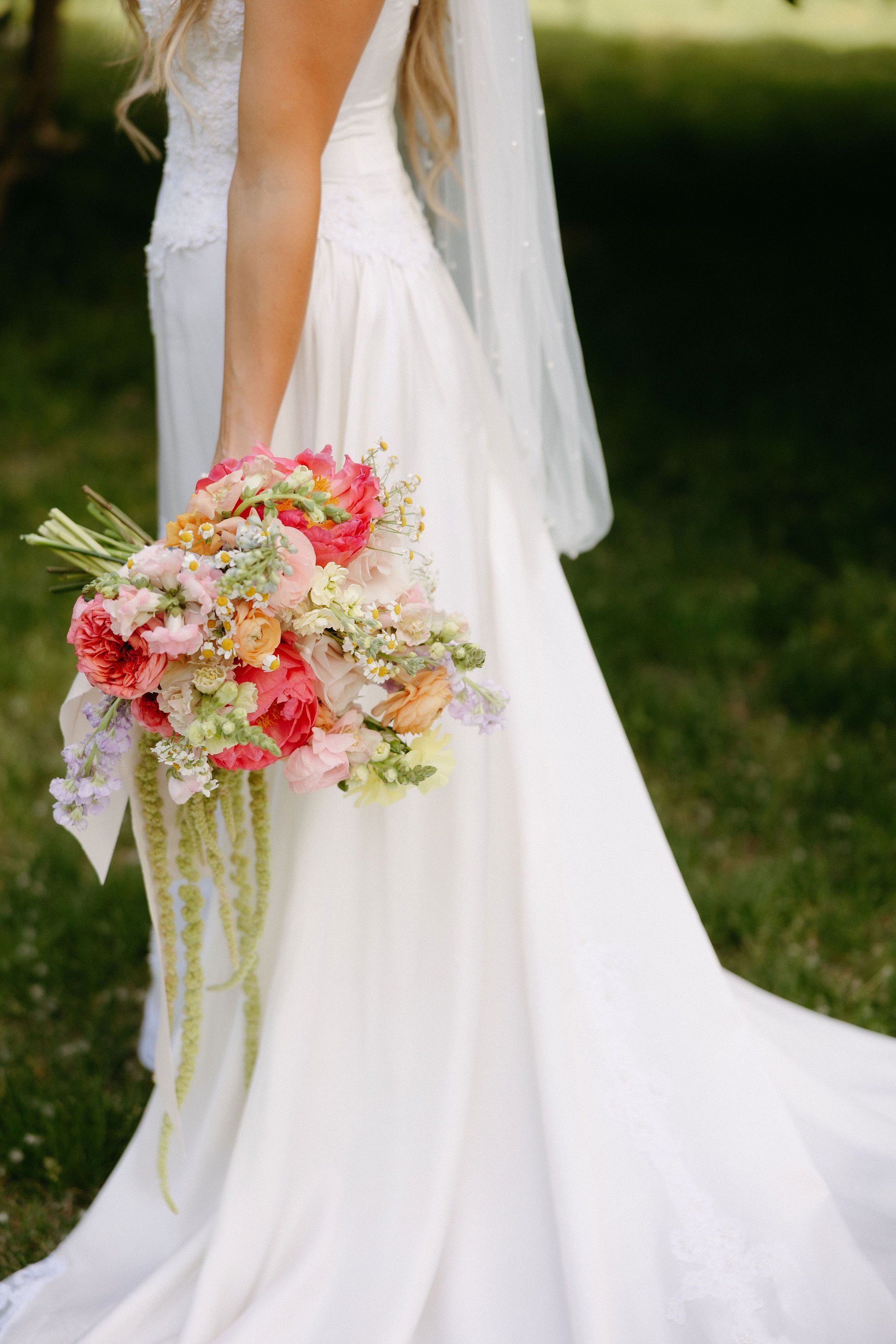 A bride in a white wedding dress holding a colorful bouquet of flowers outdoors on green grass.
