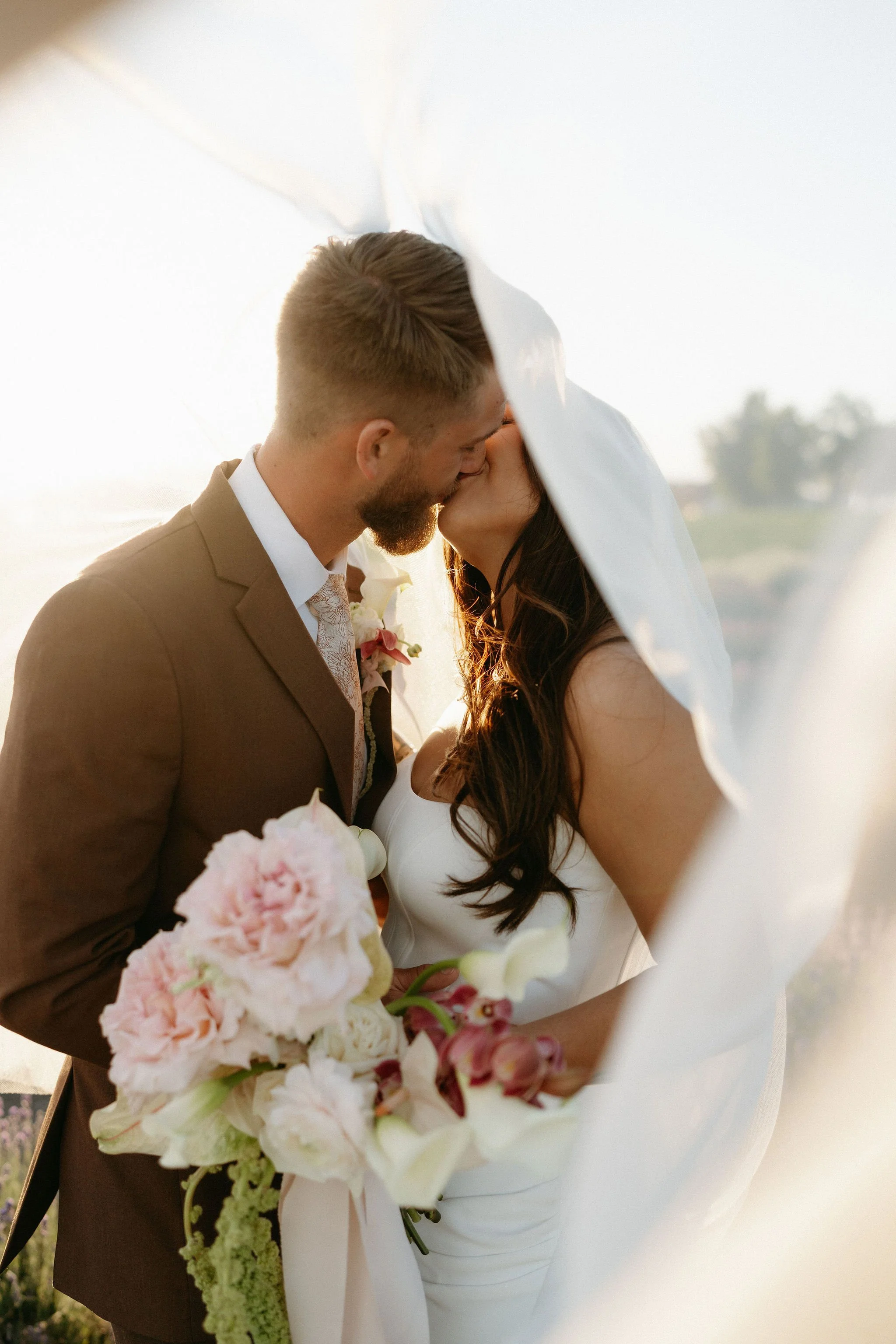 A couple sharing a kiss, likely at a wedding, with the woman holding a bouquet of pink and white flowers.