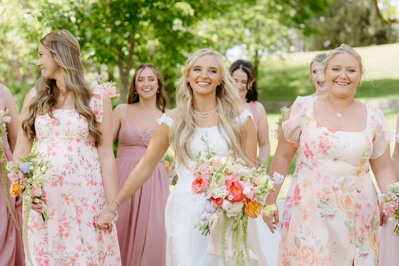 A group of women, including a bride in a white wedding dress holding a colorful bouquet, walking outdoors with friends dressed in pink floral and pastel dresses, smiling and holding hands in a park with green trees.