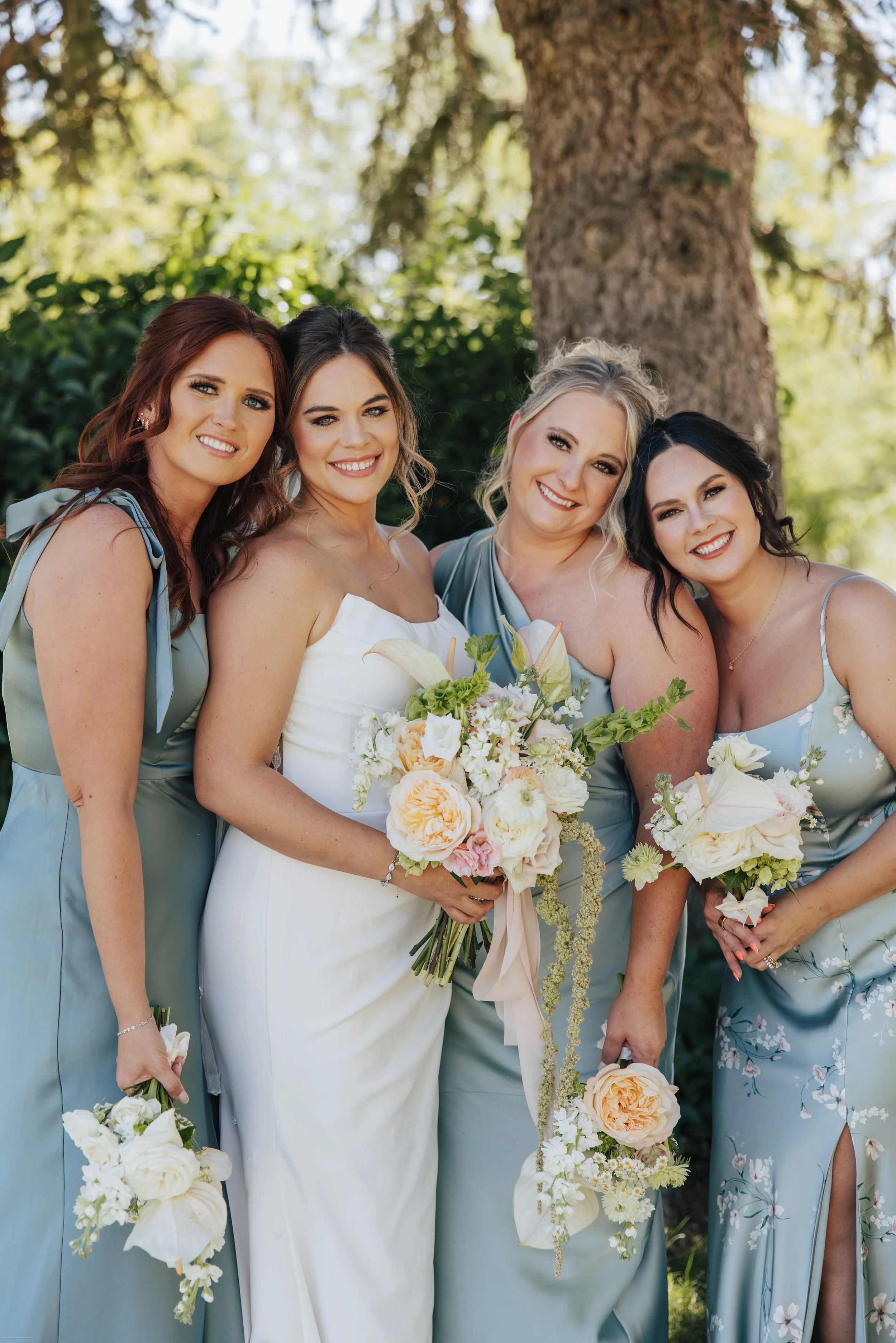 Group of smiling women in dresses holding bouquets of flowers outdoors in front of a large tree.
