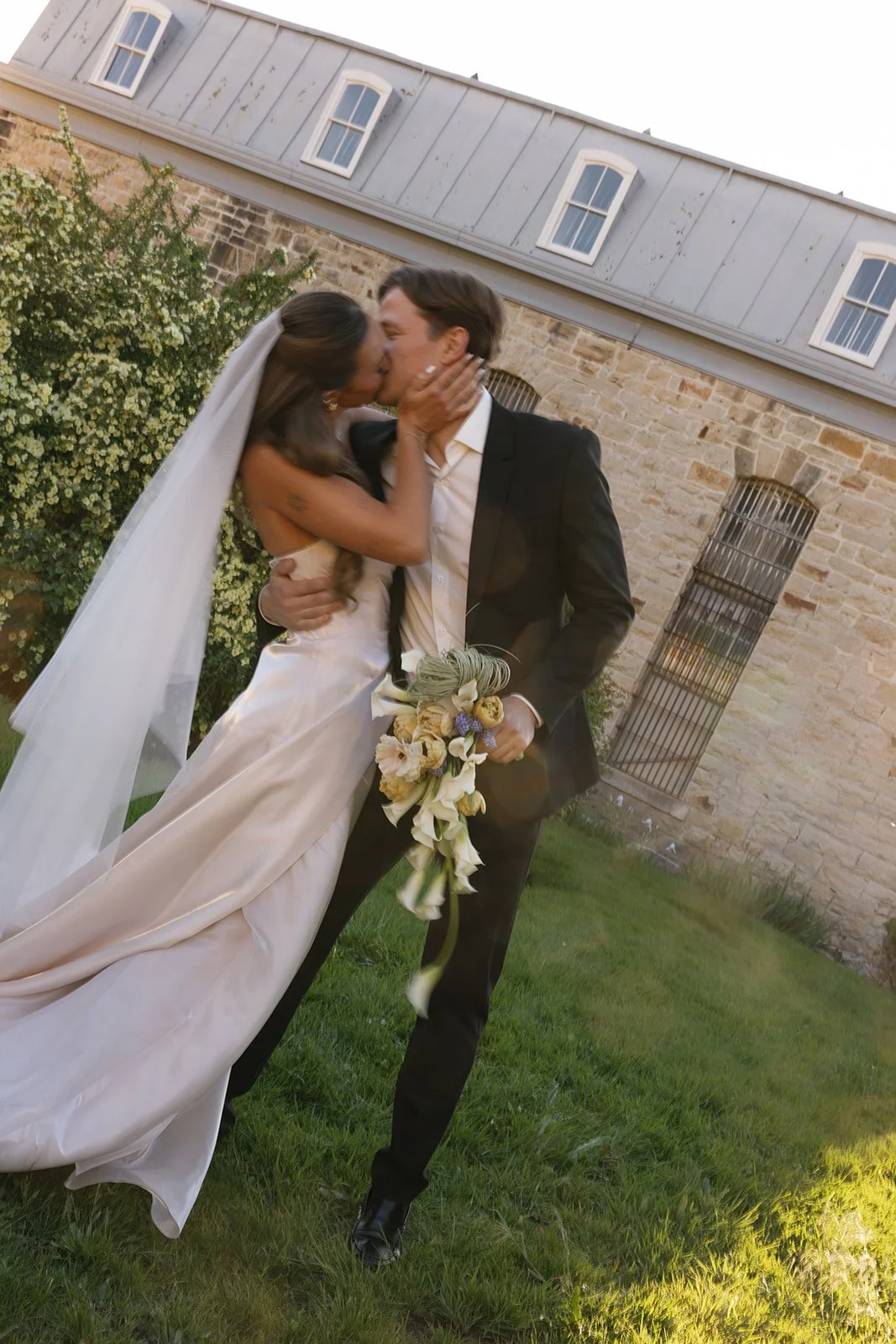 A bride and groom sharing a kiss outside on grass, with the groom holding a bouquet of flowers, in front of a stone building with windows.