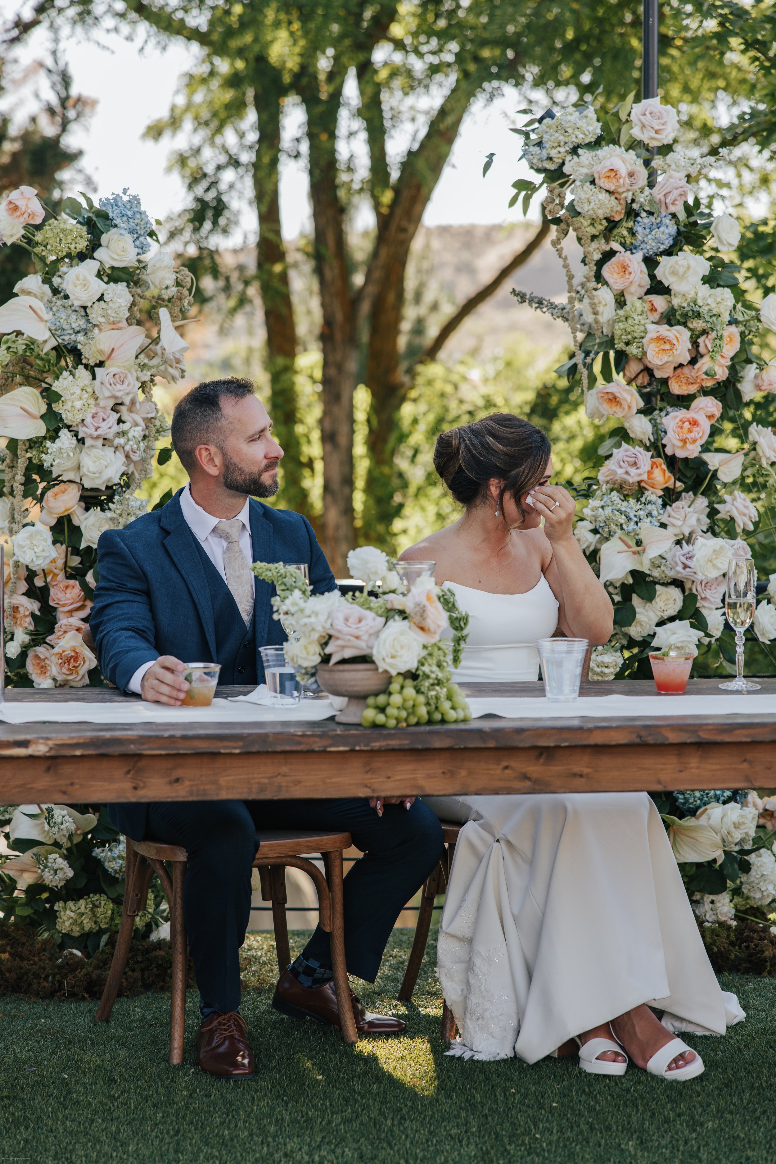 A newlywed couple sitting at a decorated table outdoors, with a floral arch behind them, during a wedding celebration. The groom is in a blue suit, and the bride is in a white wedding dress, wiping tears from her eyes.