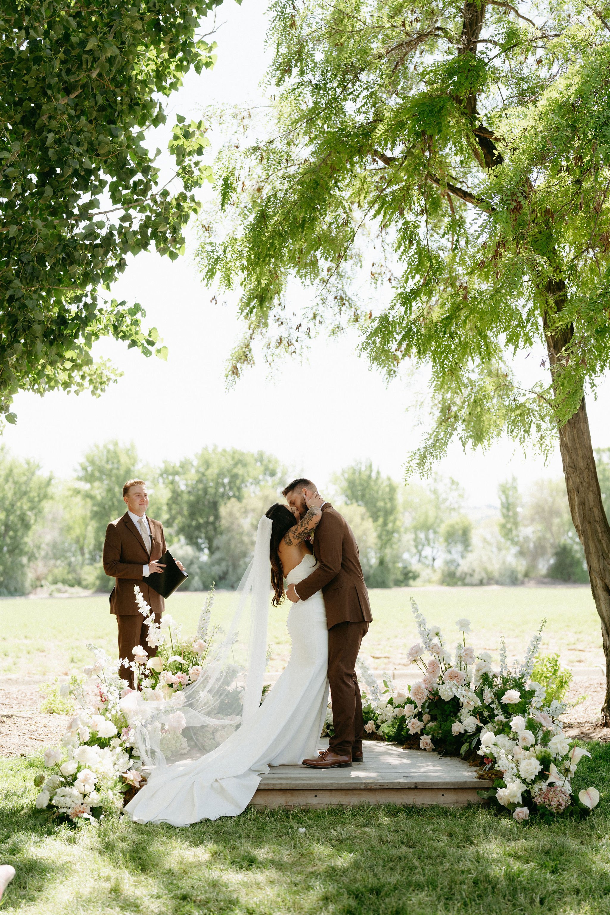 A couple shares a kiss during a wedding ceremony outdoors under a large tree, with an officiant standing nearby and floral arrangements surrounding the wooden platform.