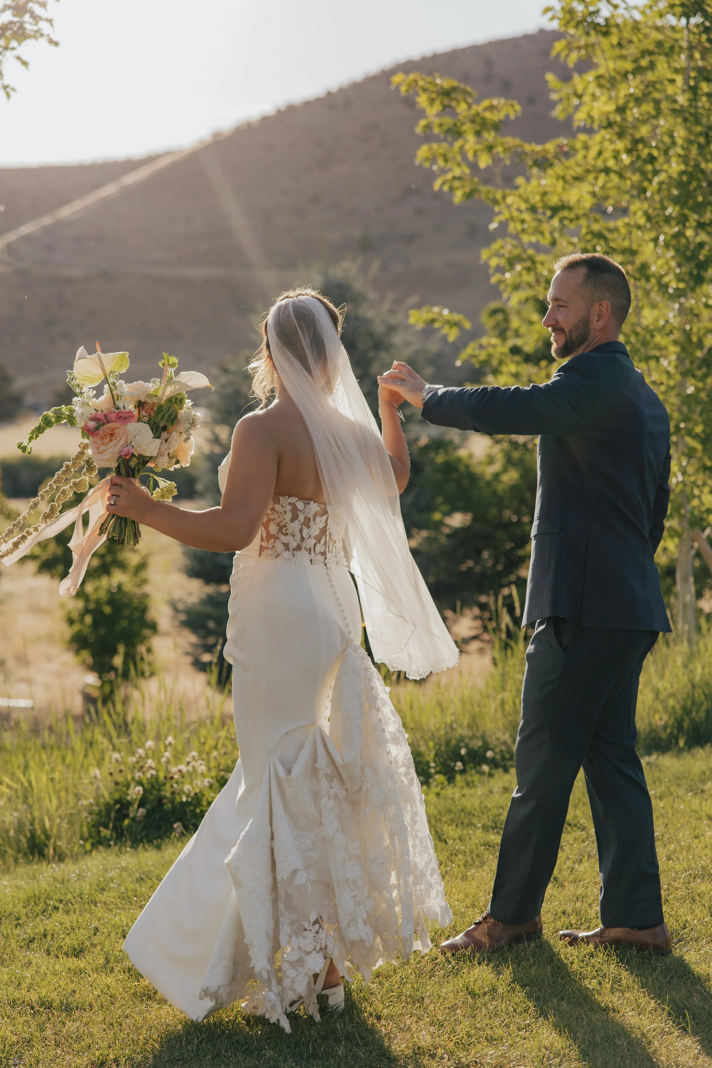 A bride and groom dancing outdoors during sunset, with the bride holding a bouquet of flowers and the groom wearing a dark suit, in a natural setting with trees and a mountain in the background.