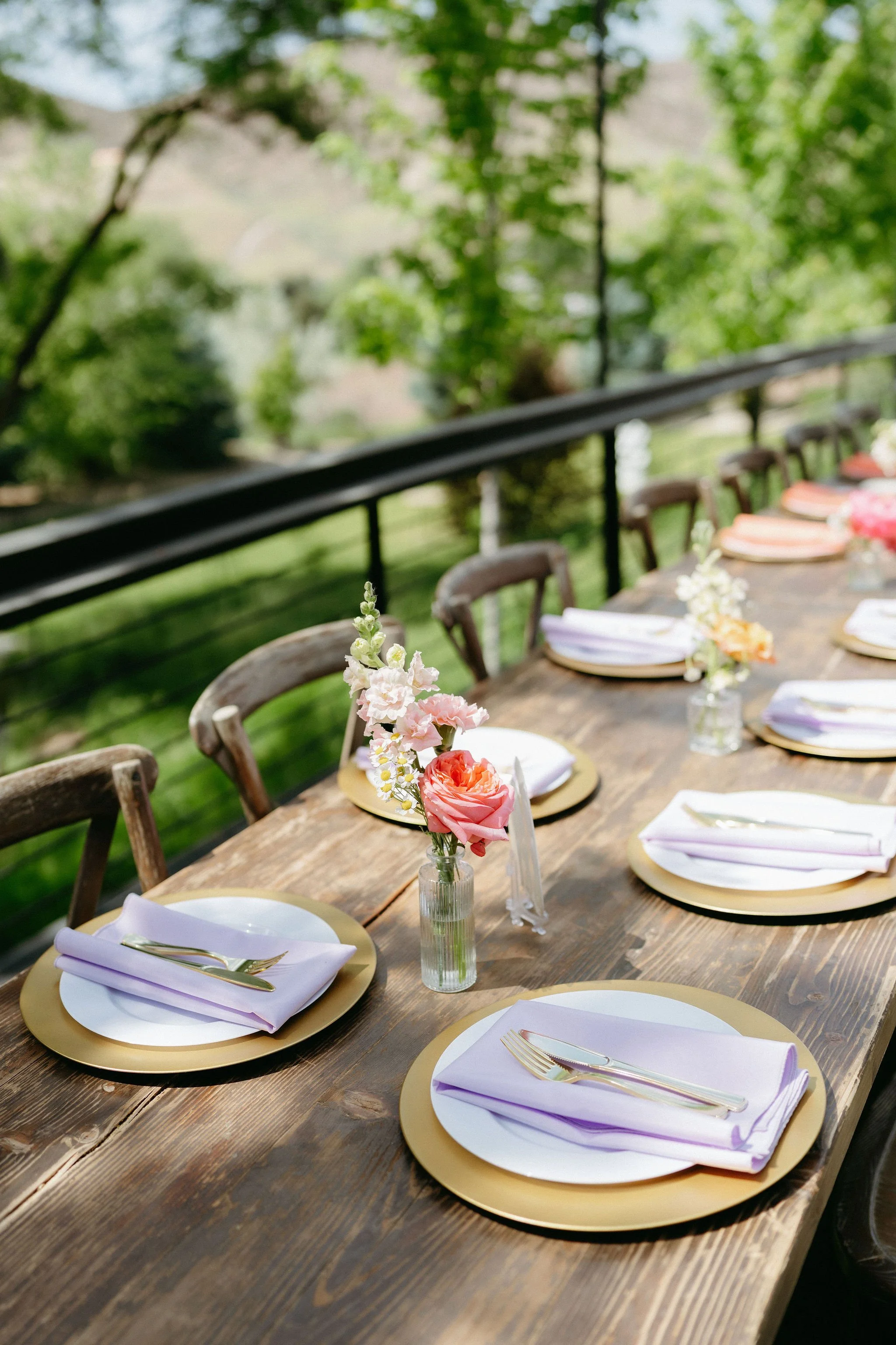 An outdoor dining table decorated for a special event with floral arrangements and tableware, surrounded by wooden chairs and lush green trees in the background.