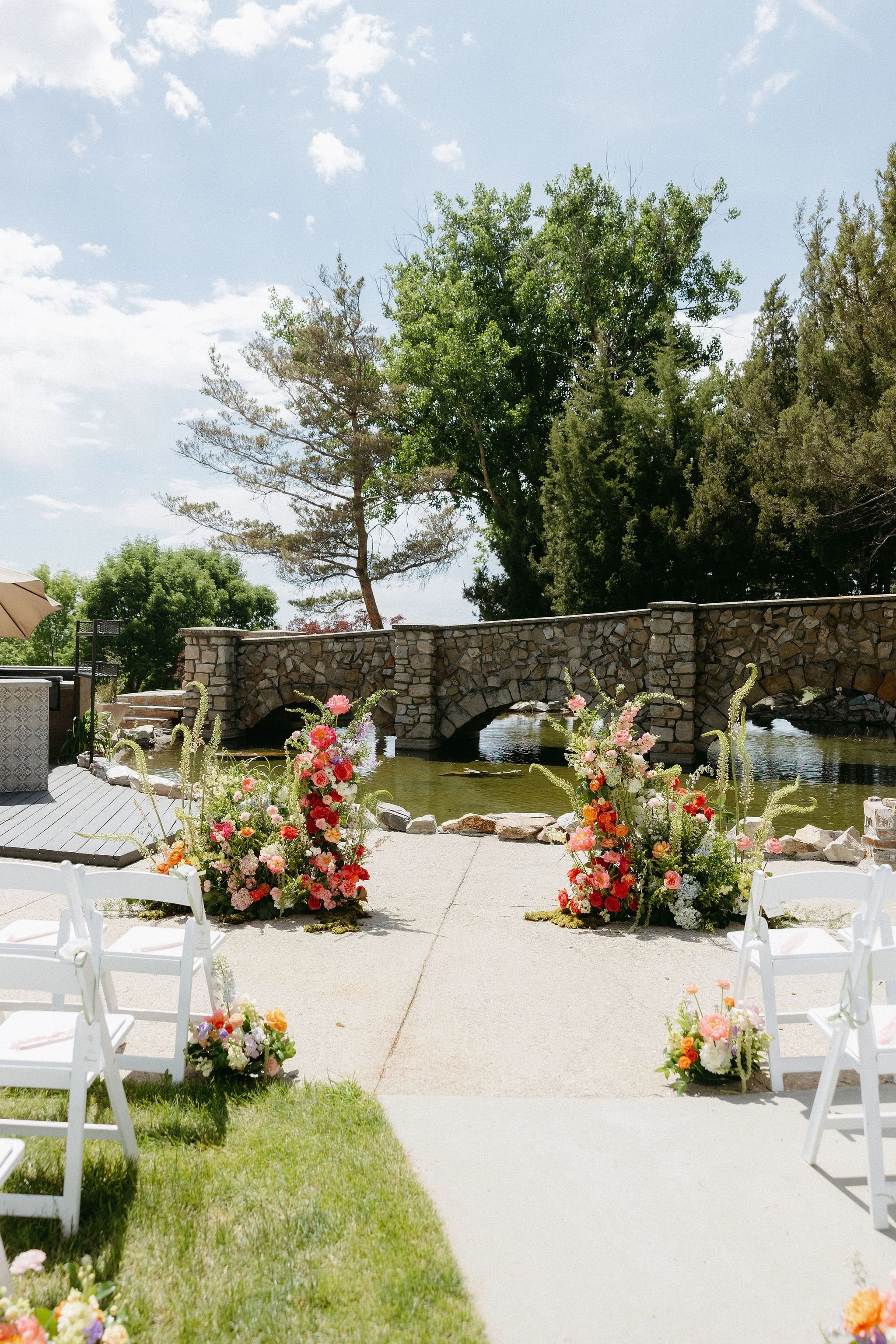 Outdoor wedding ceremony setup with white chairs, floral arrangements, a stone bridge over water, and trees in the background under a partly cloudy sky.