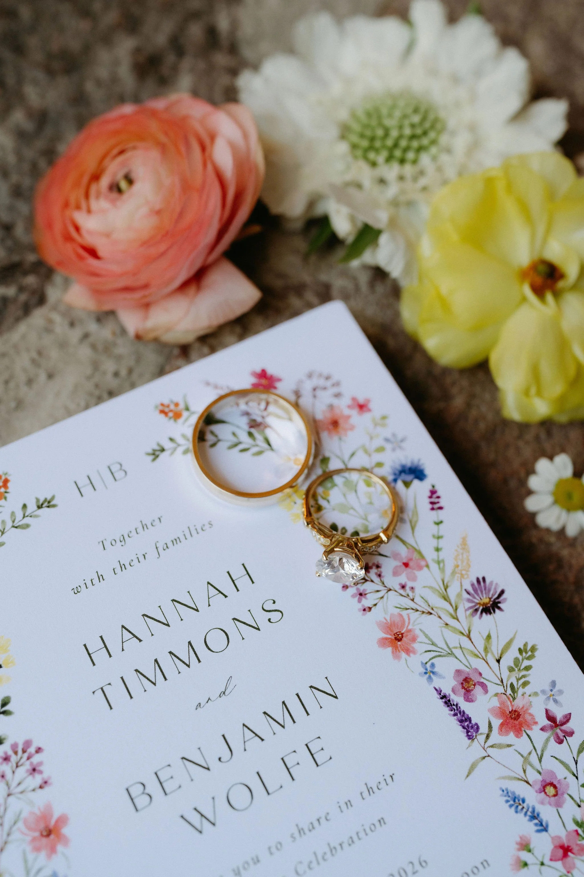 Close-up of wedding invitation card with floral border, gold wedding rings, and white and yellow flowers on a textured surface.