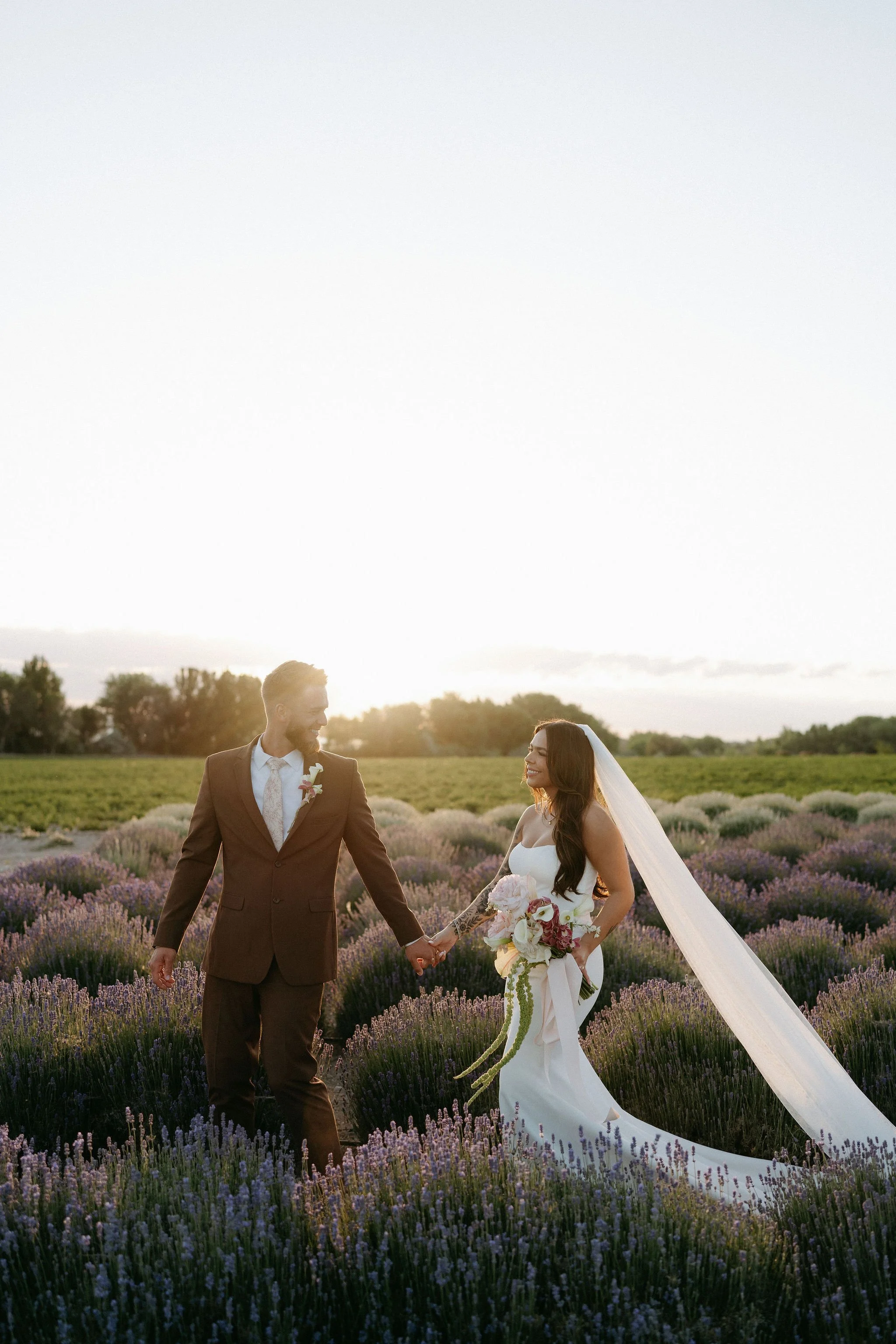 A bride and groom holding hands in a lavender field during sunset. The bride is in a white wedding dress with a long veil and is holding a bouquet, while the groom is in a brown suit with a pink boutonniere. They are smiling and looking at each other