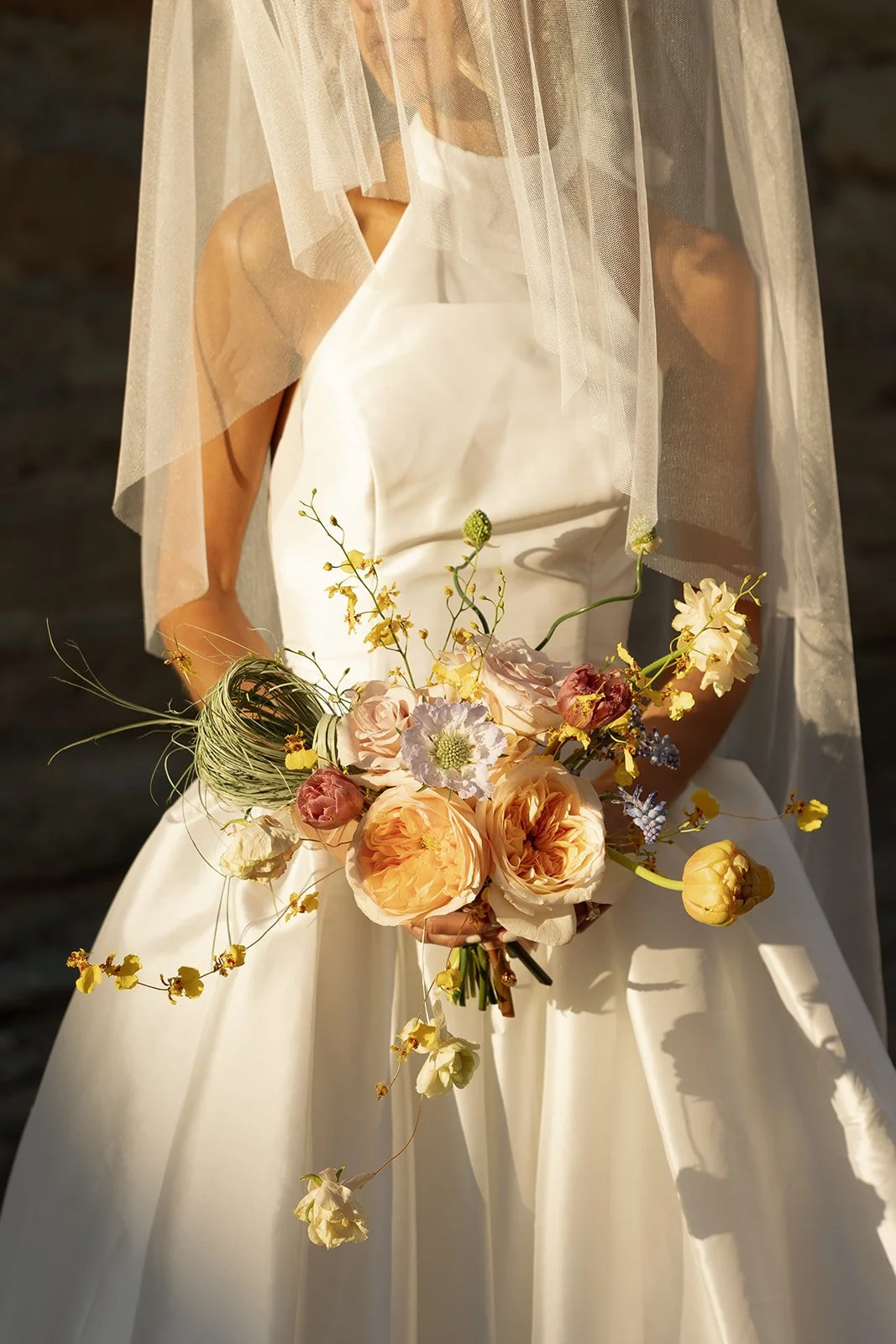 A bride in a white satin wedding dress holding a bouquet of pastel-colored flowers, with a sheer veil over her face, standing outdoors.