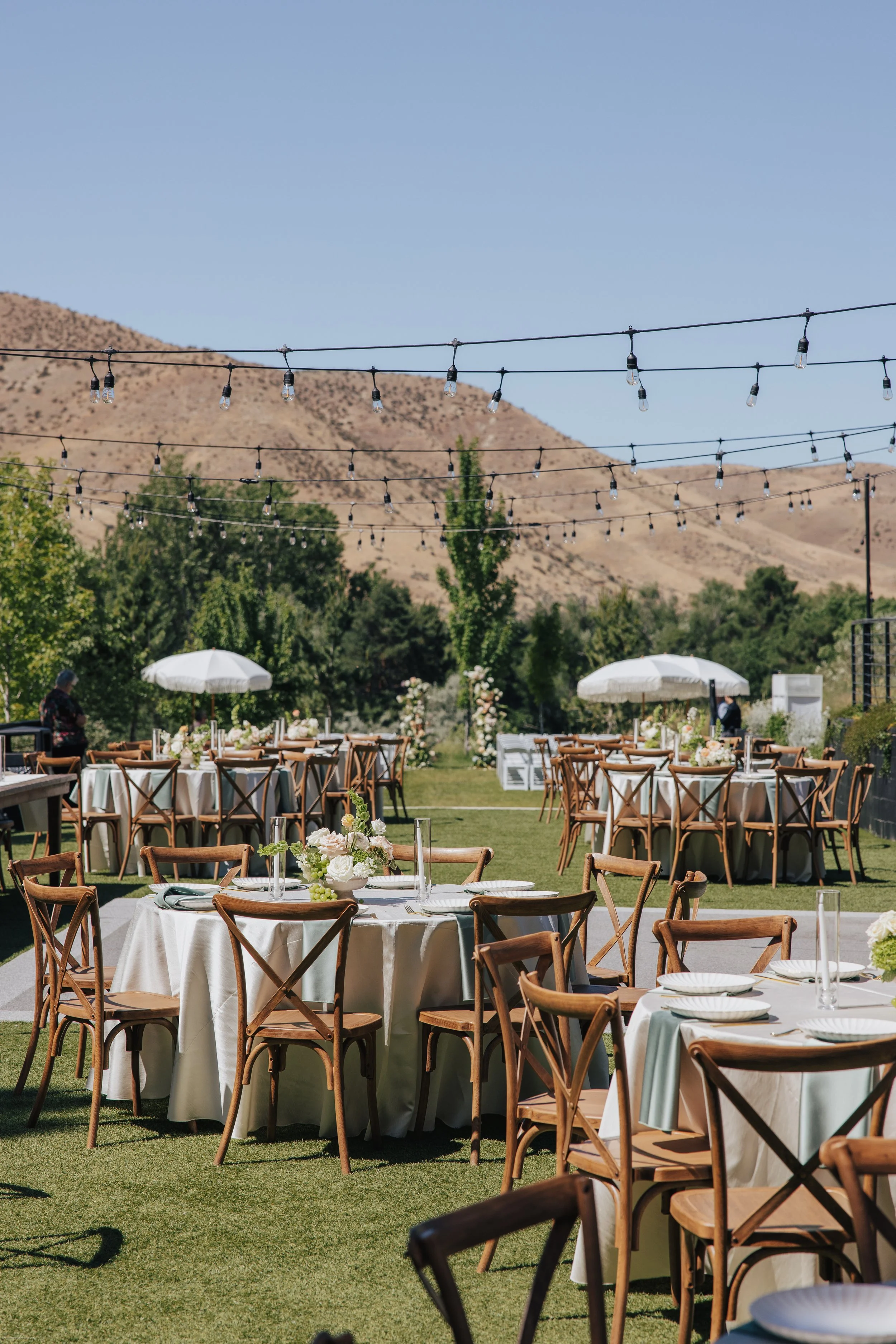 Outdoor celebration setup with tables and chairs, decorated with flowers, under string lights, with a backdrop of mountains and trees.