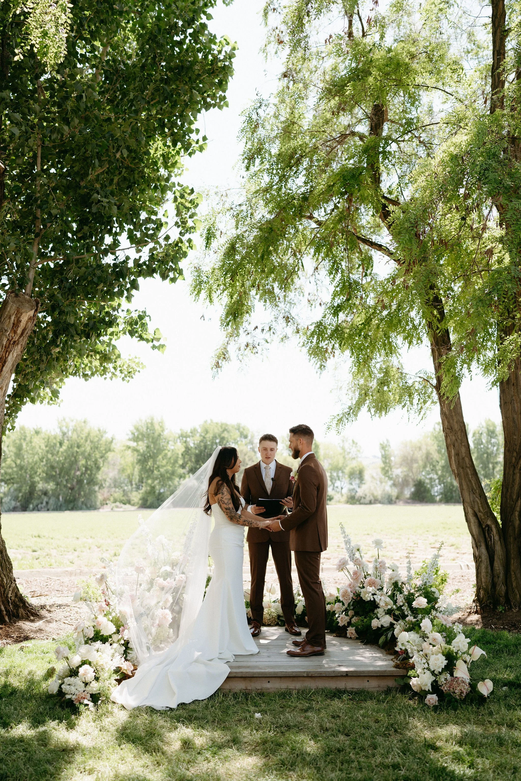A couple getting married outdoors under tall trees, with an officiant standing in front of them. The bride is in a white wedding dress with a long veil and tattoos on her arm. The groom is in a brown suit. Decorations include white and pink flowers w