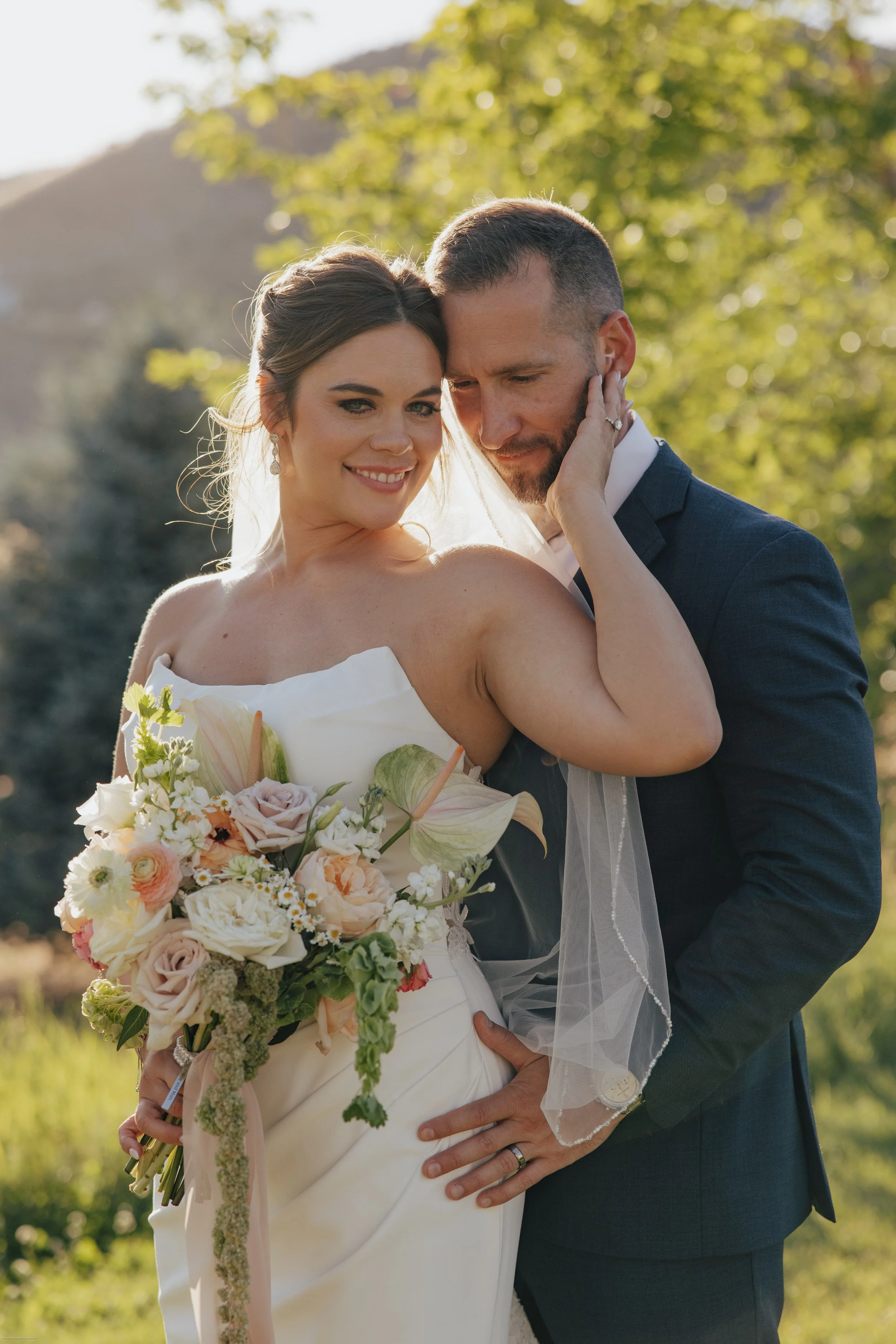 A bride and groom are embracing during their outdoor wedding, with greenery and sunlight in the background. The bride holds a large bouquet of pastel-colored flowers.