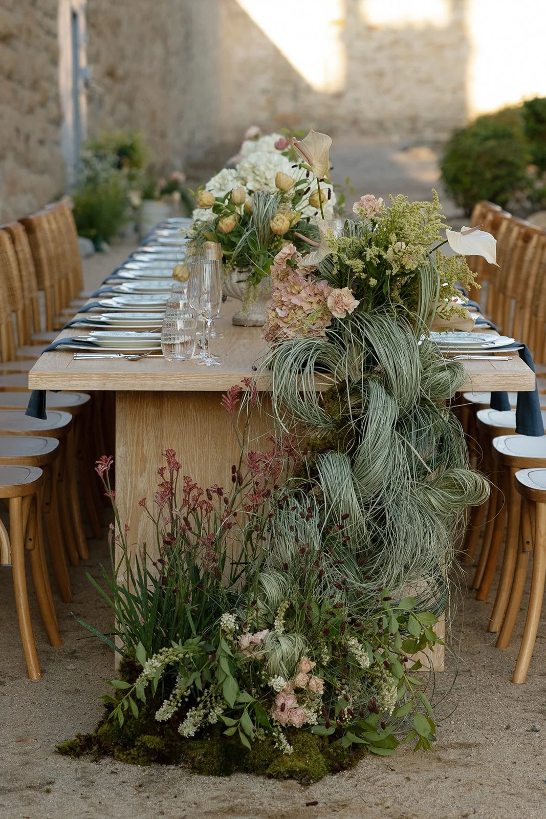 A long wooden dining table decorated with floral centerpieces, set outdoors against a stone wall. The table has wine glasses, plates, and utensils, with chairs on either side. The floral arrangements include white, pink, and green flowers and greenery, cascading down the sides of the table.