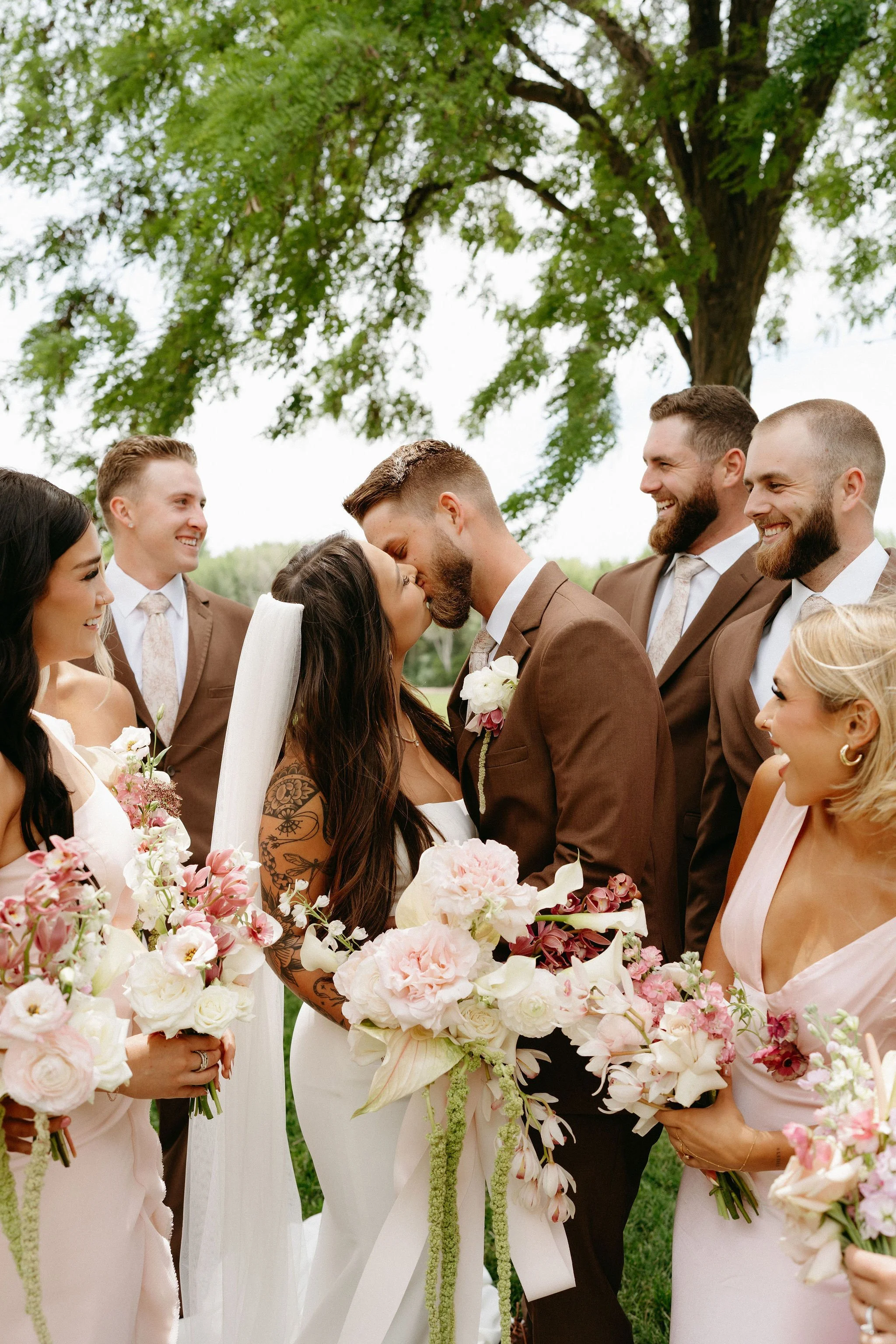 A wedding ceremony outdoors with the bride and groom kissing, surrounded by bridesmaids and groomsmen holding pink and white flowers, under a large tree.