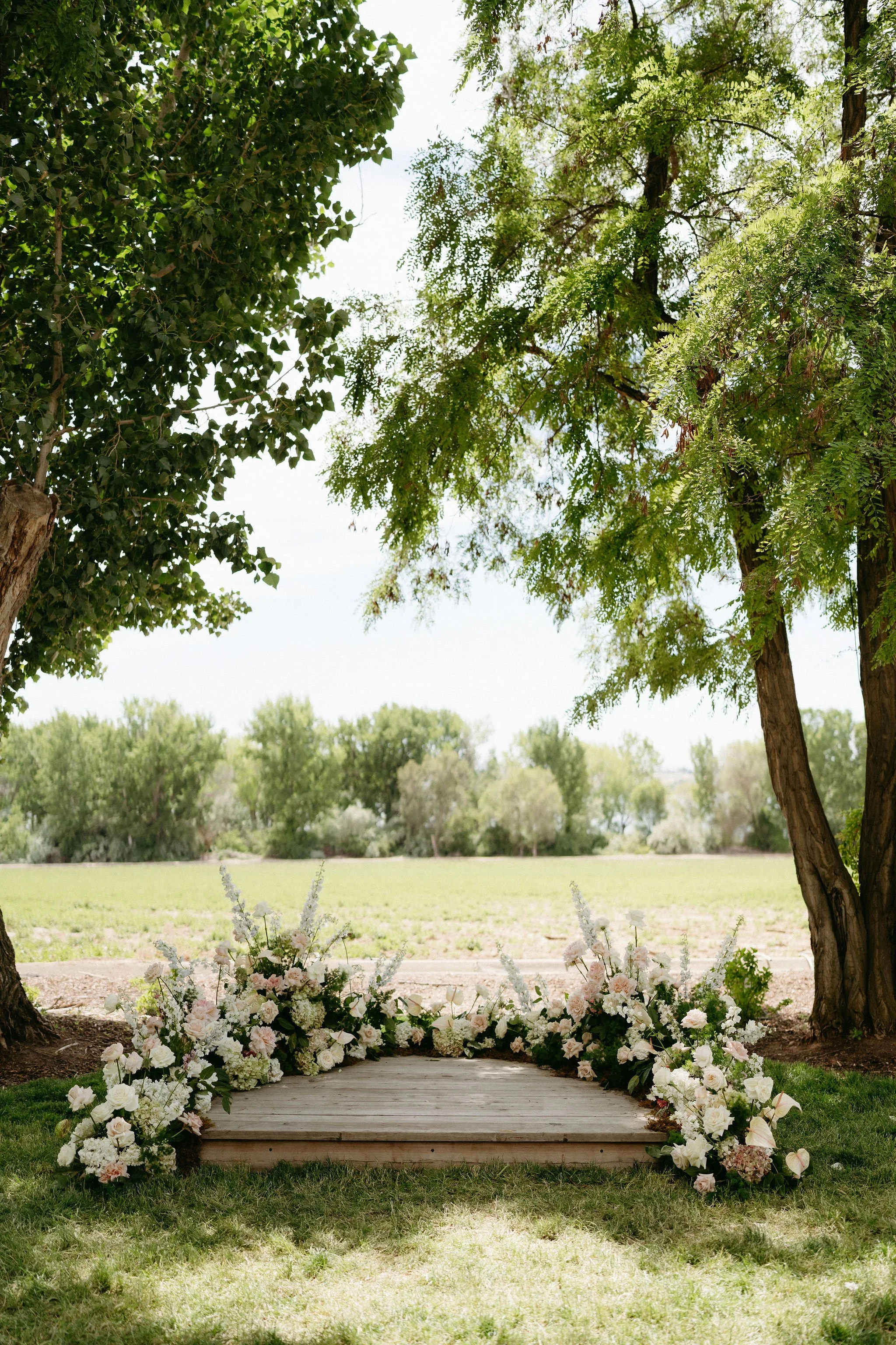 Bouquet of white and blush pink flowers arranged along a wooden pathway, with trees and greenery in the background, set outdoors for a wedding or special event.