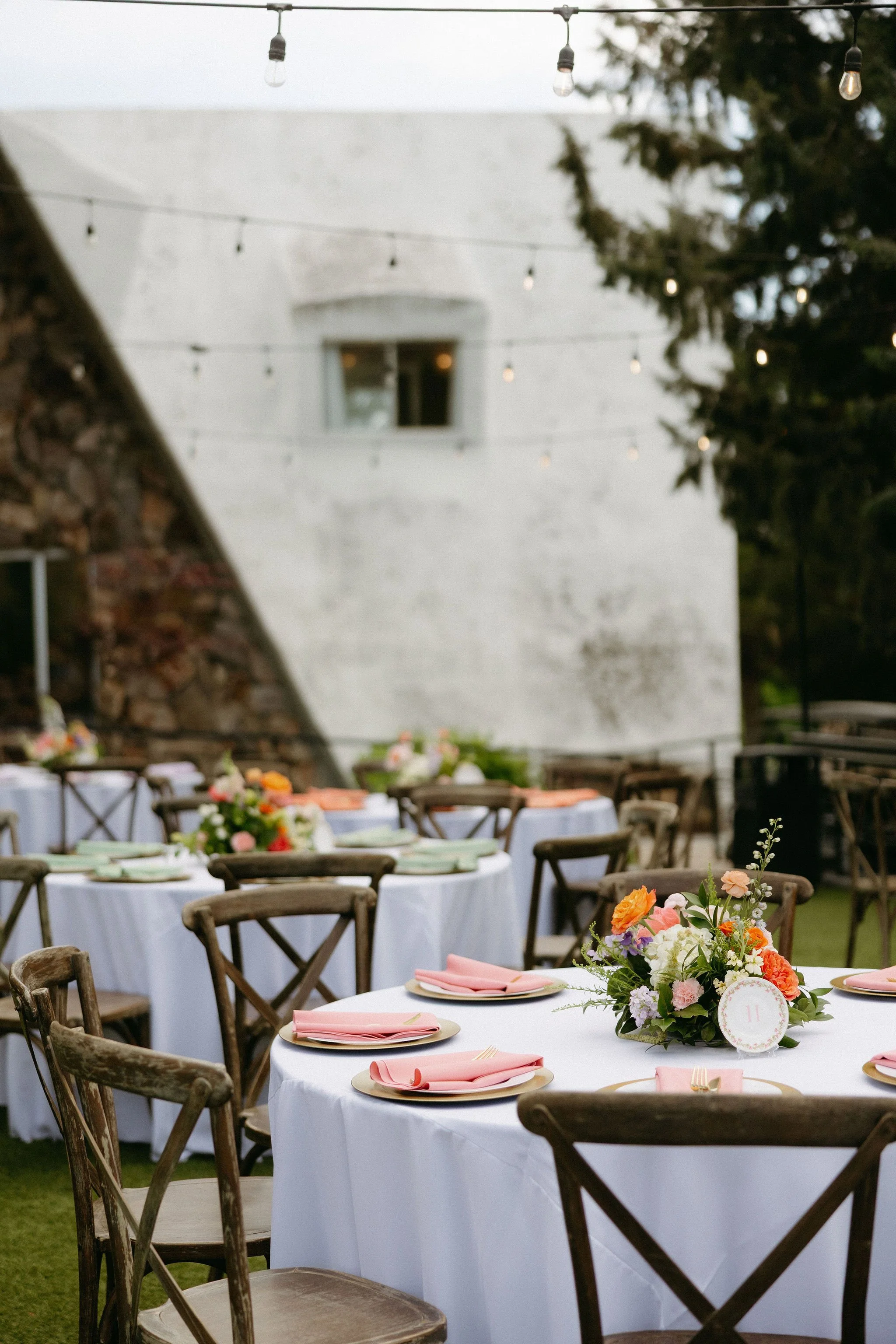 Outdoor wedding reception with round tables covered in white tablecloths, decorated with floral centerpieces, pink napkins, and gold plates. Wooden chairs surround the tables. String lights hang above, and a white building with a small window is in t