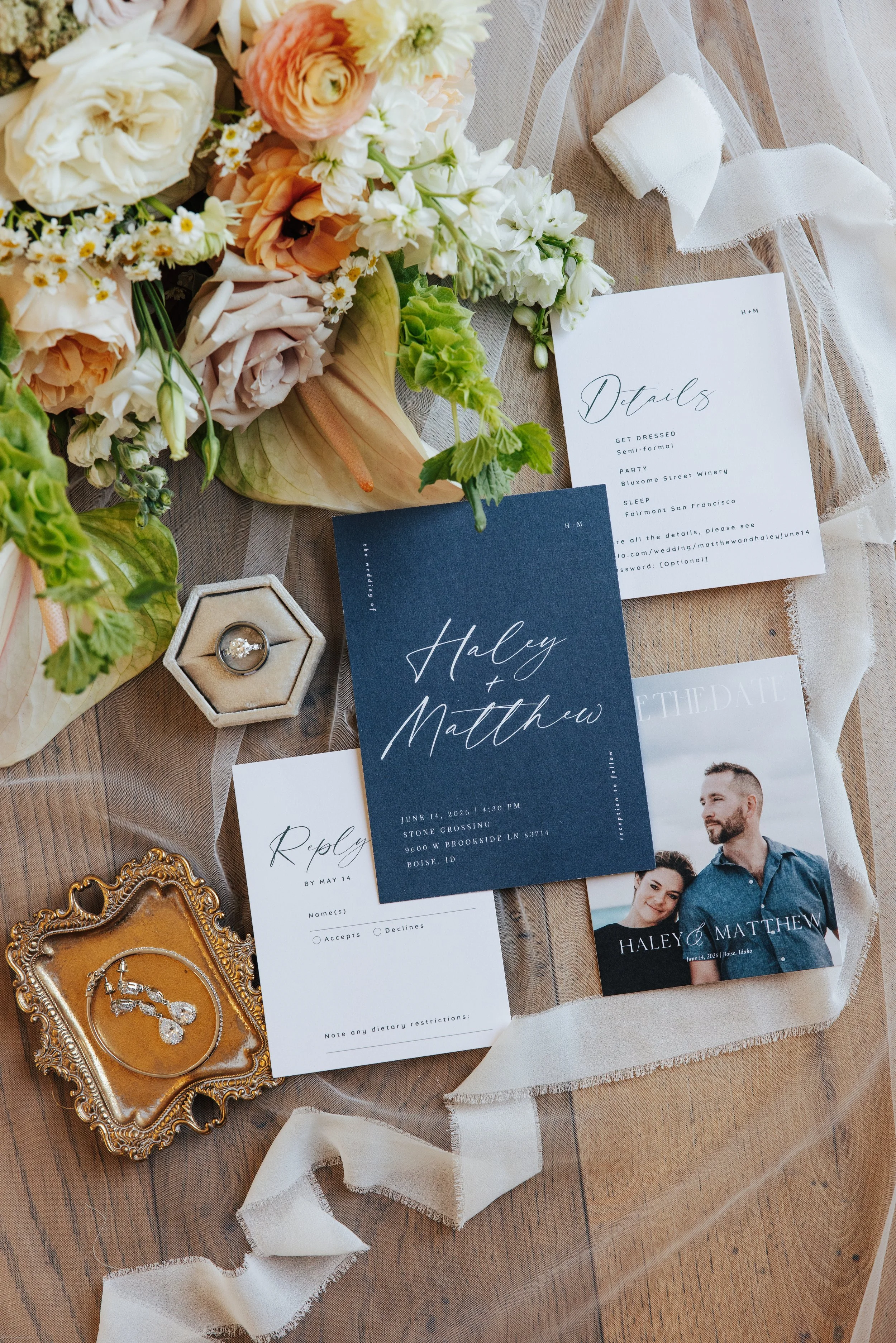 Wedding invitations, floral bouquet, a ring in a decorative box, and other wedding details on a wooden table with a light fabric.