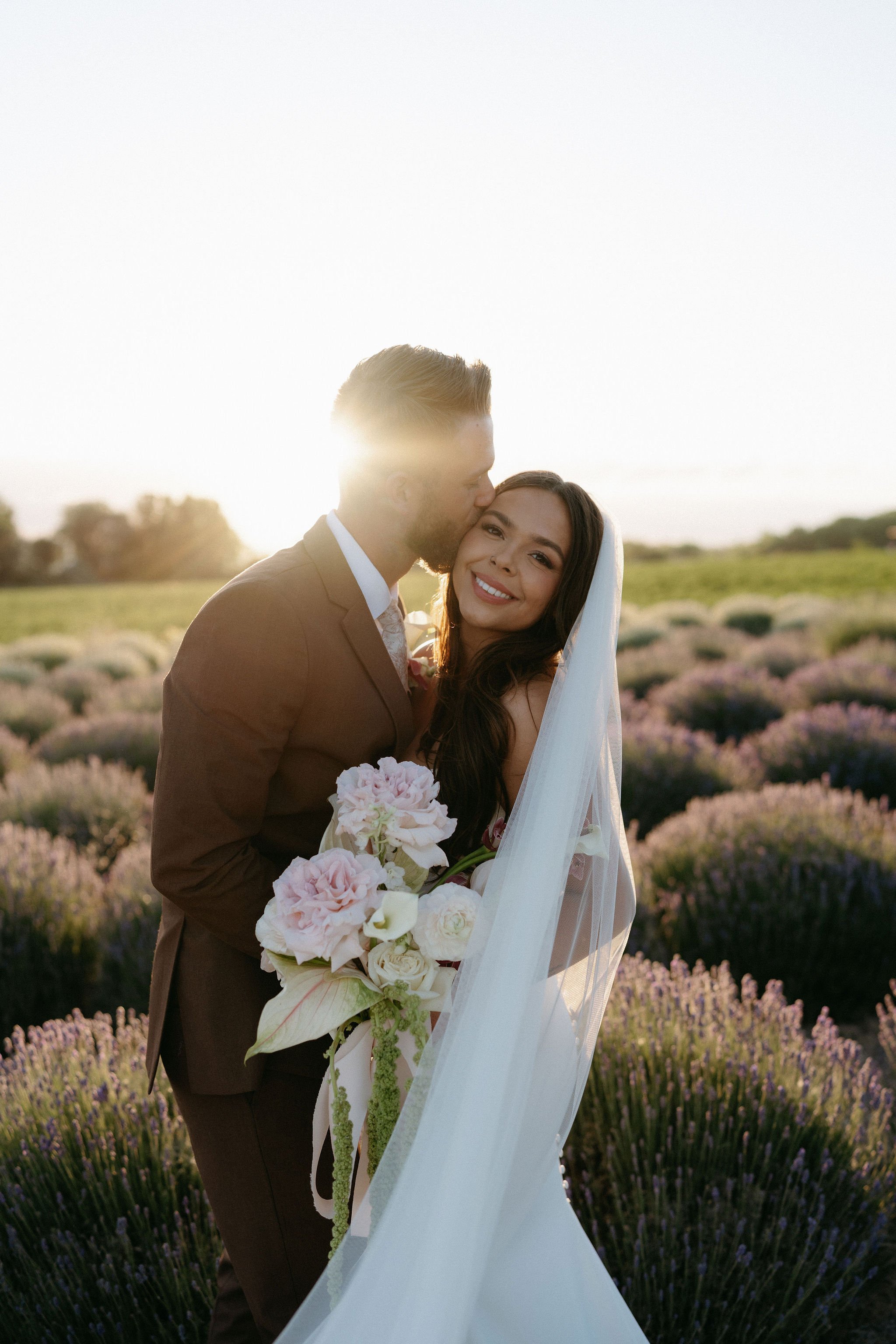 A newlywed couple standing in a lavender field at sunset. The groom, dressed in a brown suit, kisses the bride on the forehead. The bride, wearing a white wedding dress and veil, holds a bouquet of pink and white flowers and smiles at the camera.