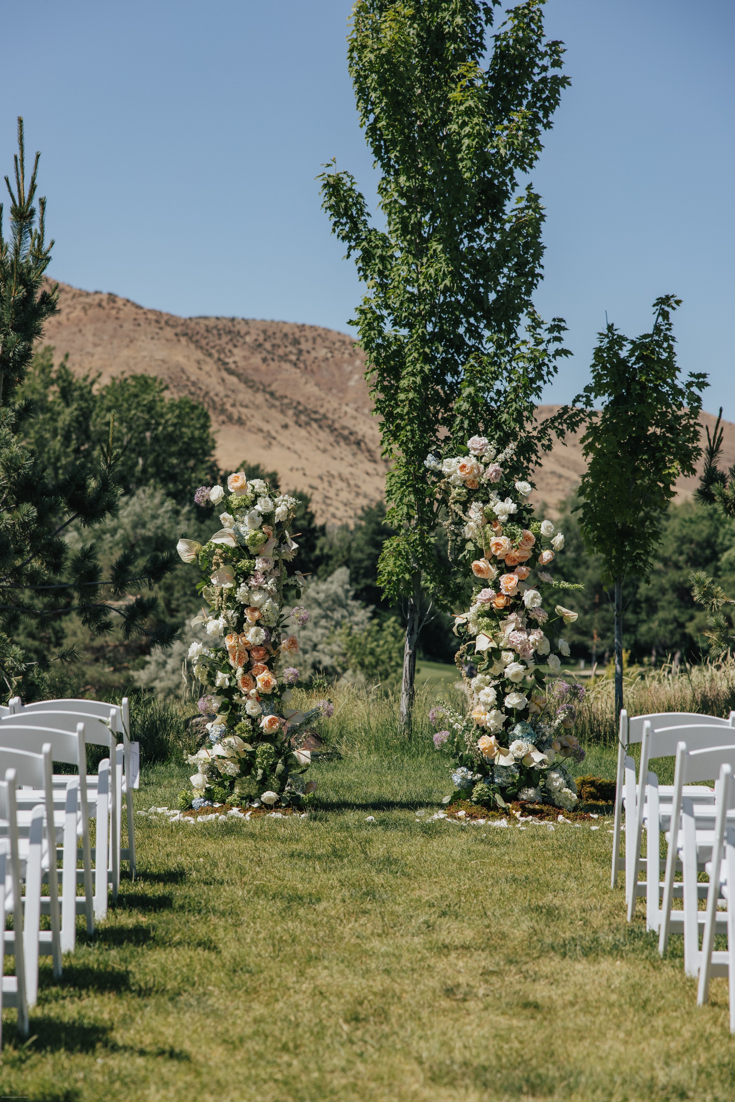 Outdoor wedding ceremony setup with white chairs arranged on both sides of an aisle, and a floral arch made of pink, white, and purple flowers, set against a backdrop of trees and mountains under a clear blue sky.