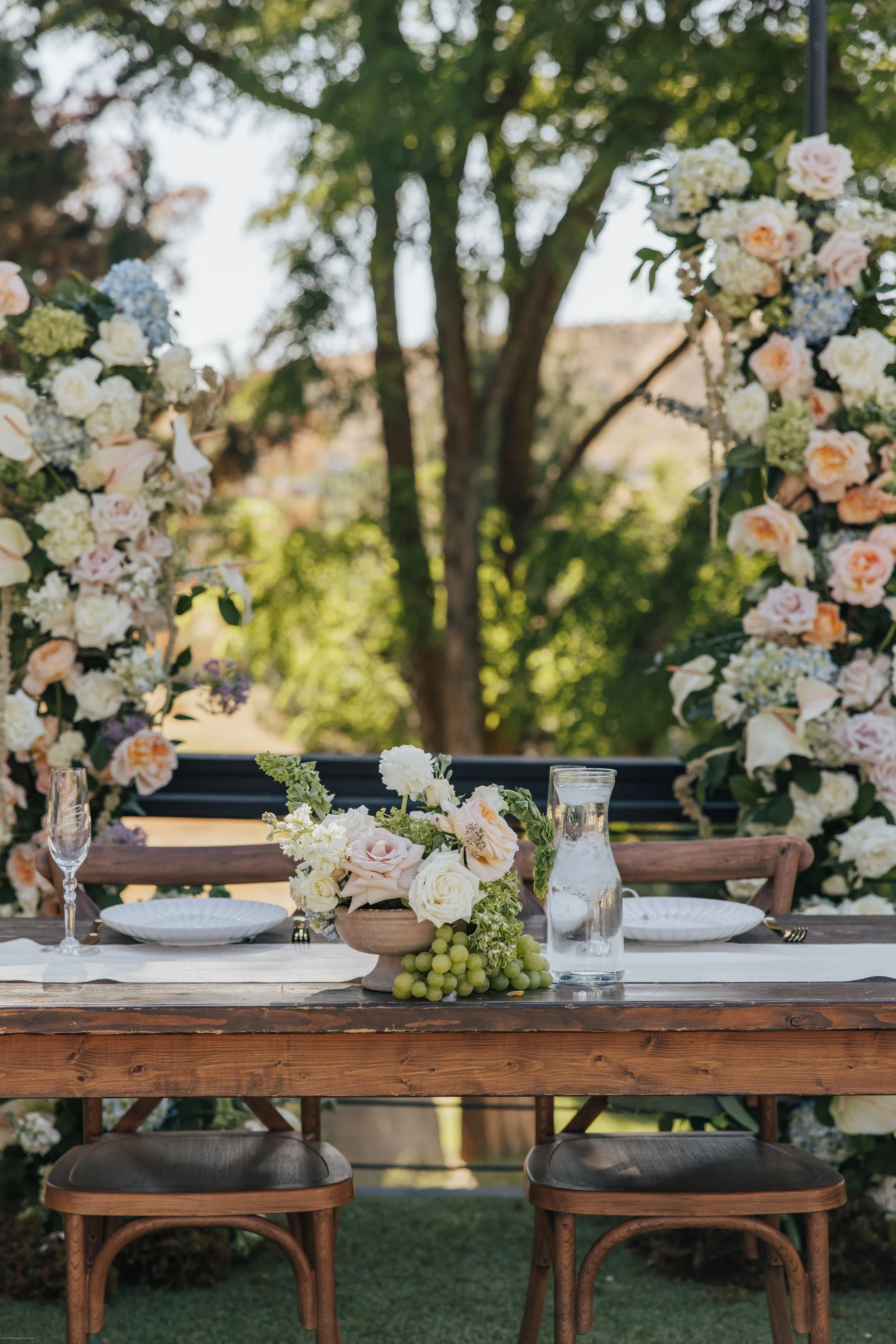 Outdoor wedding table setup with a wooden table, white plates, glasses, and a floral centerpiece featuring white and pale pink roses, grapes, and greenery, with a floral arch in the background under trees.