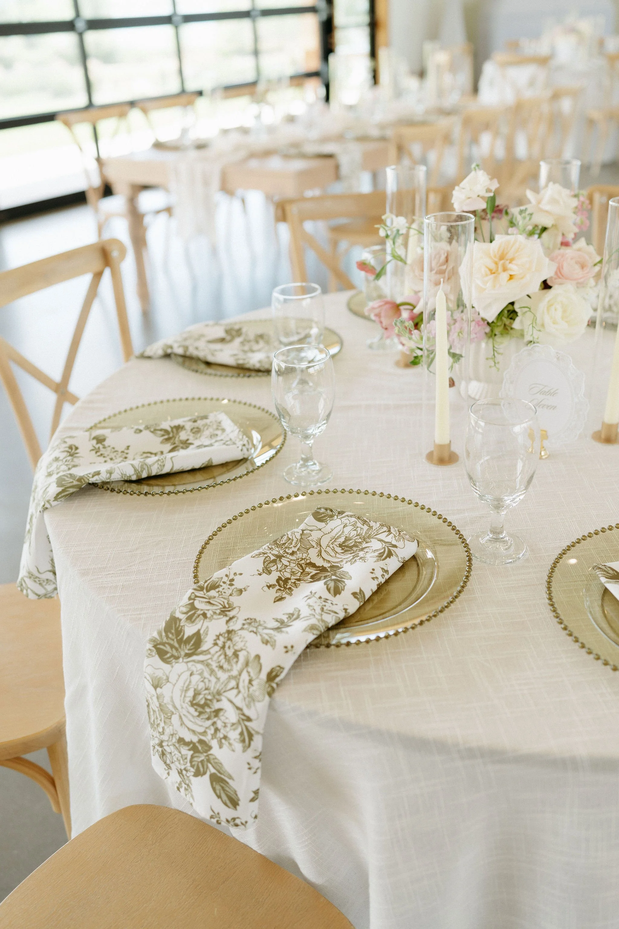 Elegant banquet table set with floral centerpiece, candles, glassware, and napkins in a bright room with large windows.