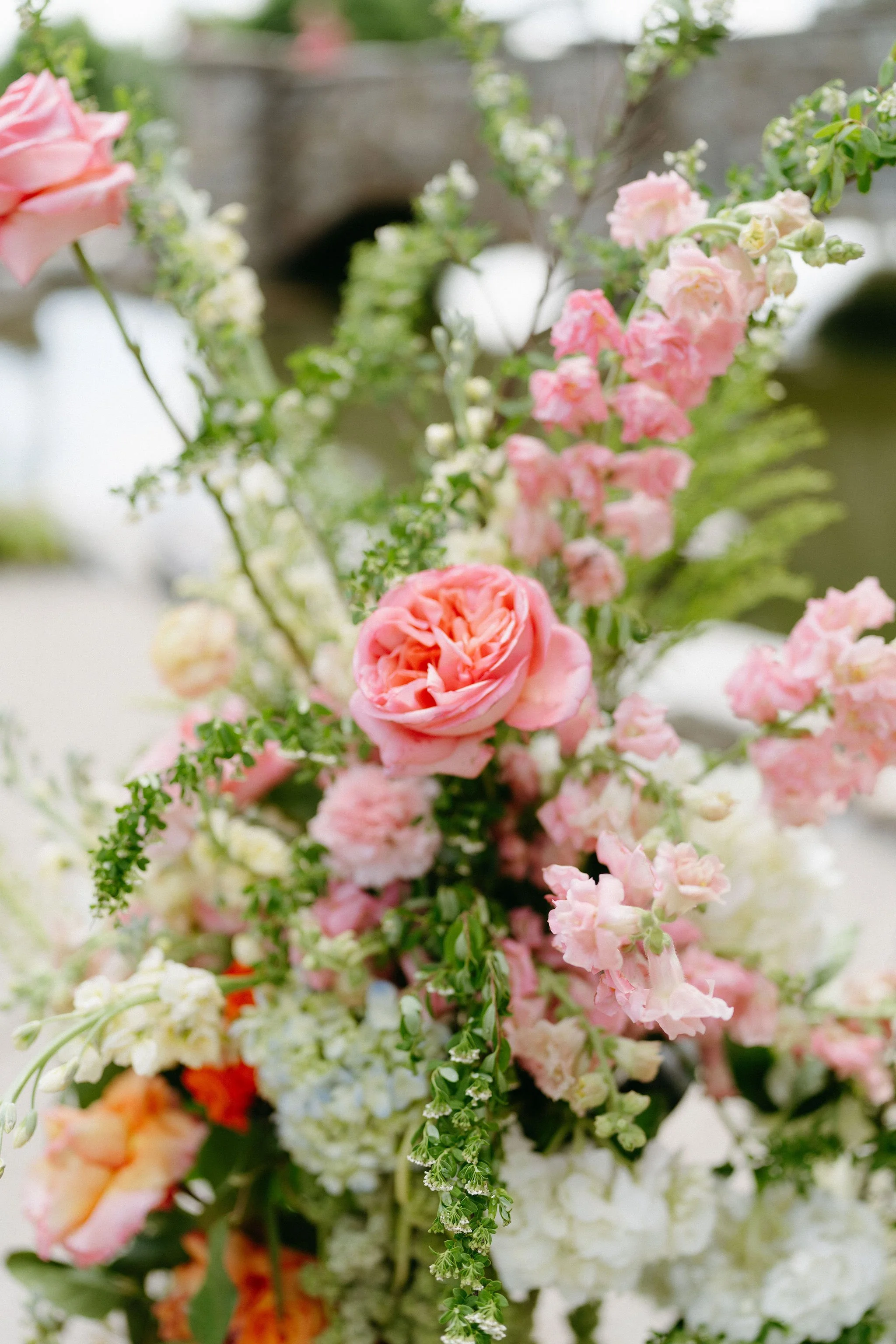 A close-up of a colorful flower arrangement with pink, white, and orange flowers, including roses and snapdragons, with blurred background.
