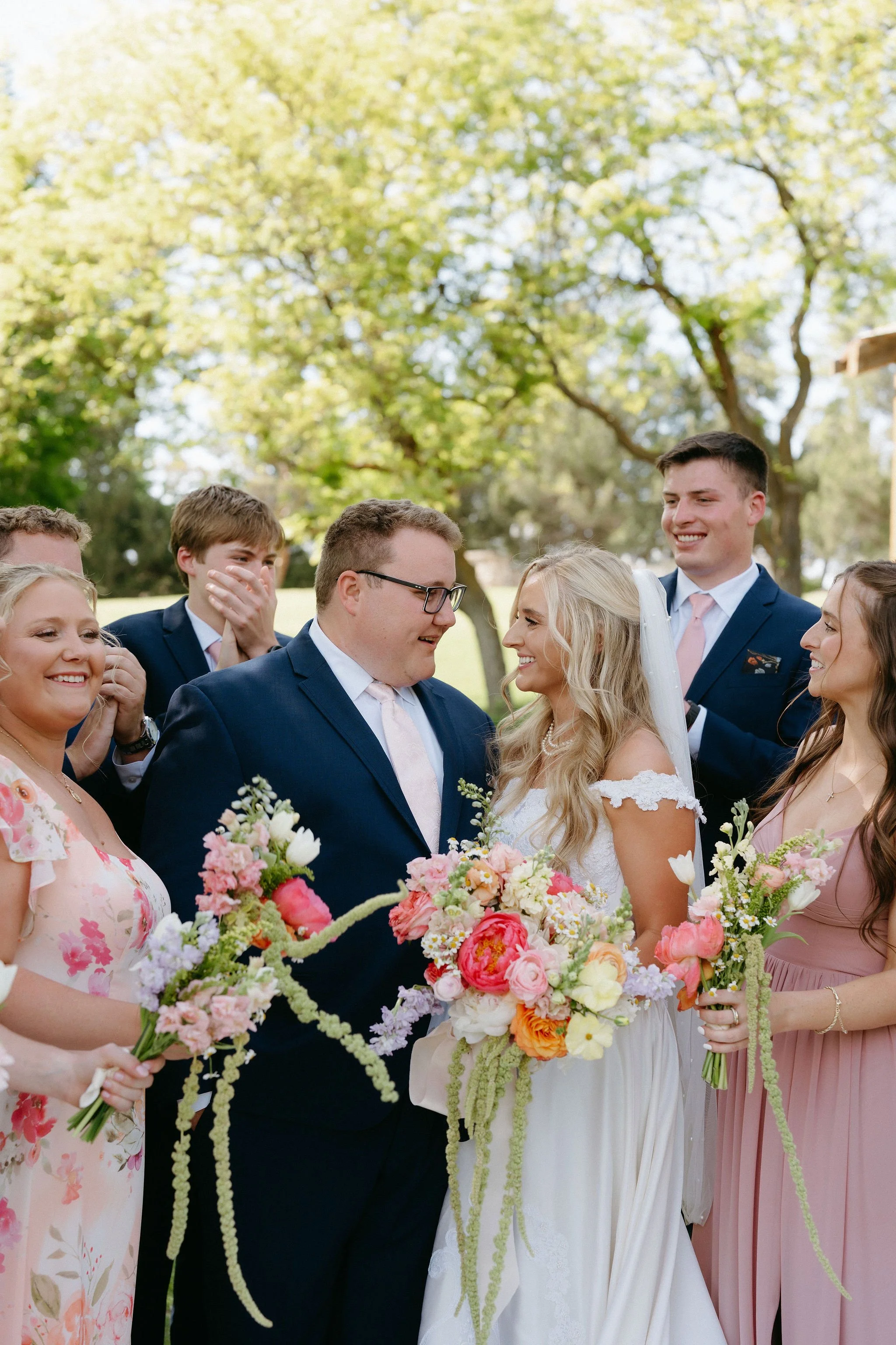 A wedding ceremony outdoors with the bride and groom standing close, surrounded by bridesmaids and groomsmen, holding bouquets, with green trees in the background.