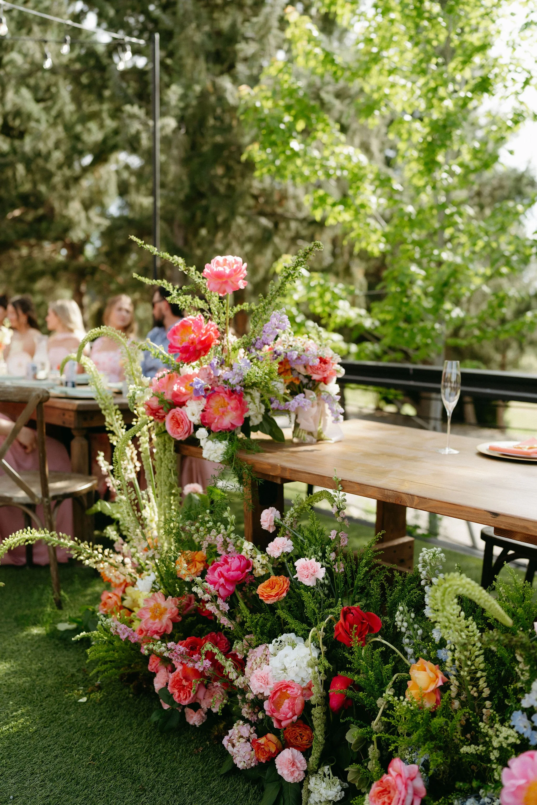 Colorful flower arrangements and bouquets placed on a wooden table outdoors, surrounded by green grass and trees, with people seated in the background at an outdoor event.