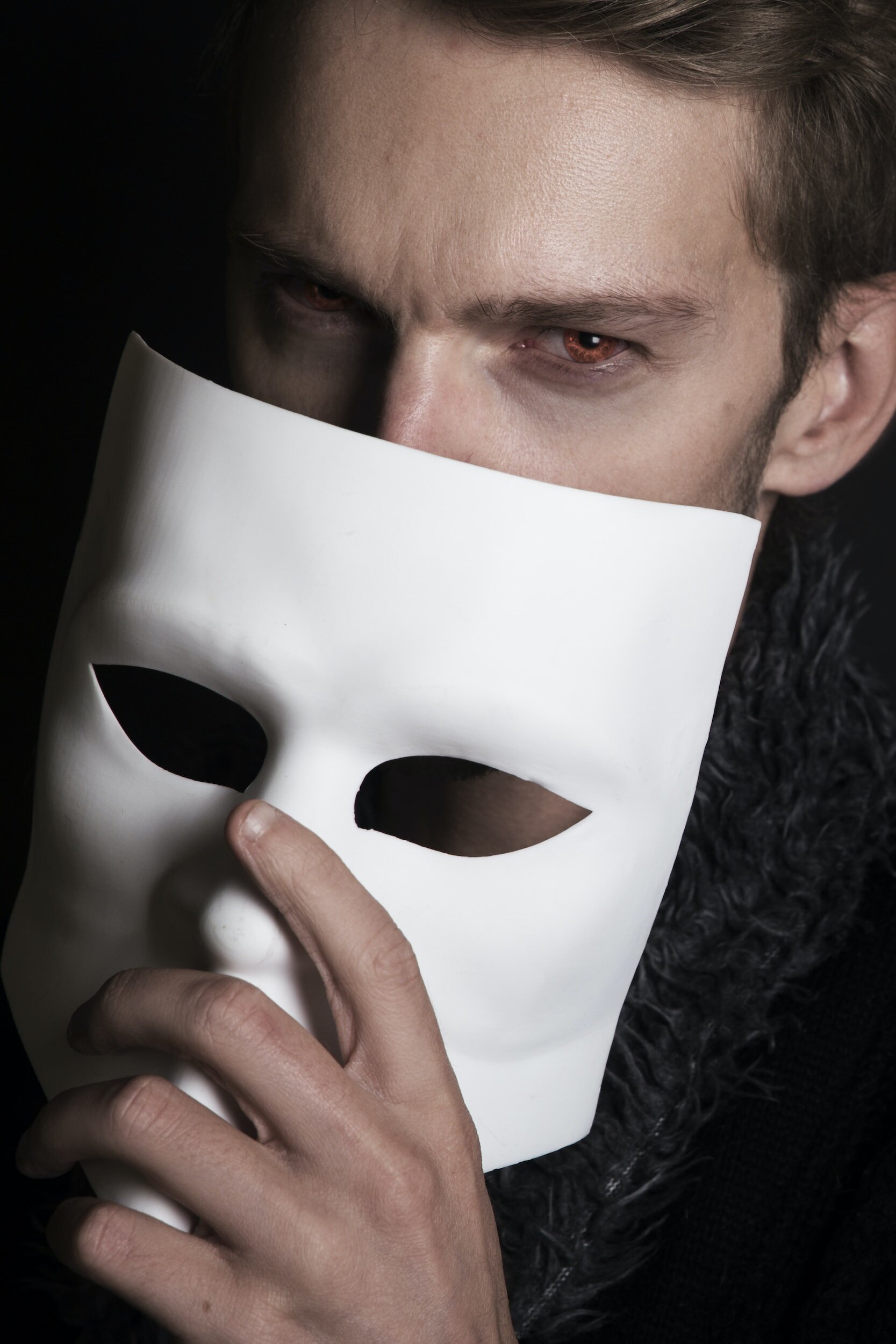 Close-up of a person holding a white mask with eye cutouts, partially covering their face, with visible intense eyes and a dark background.