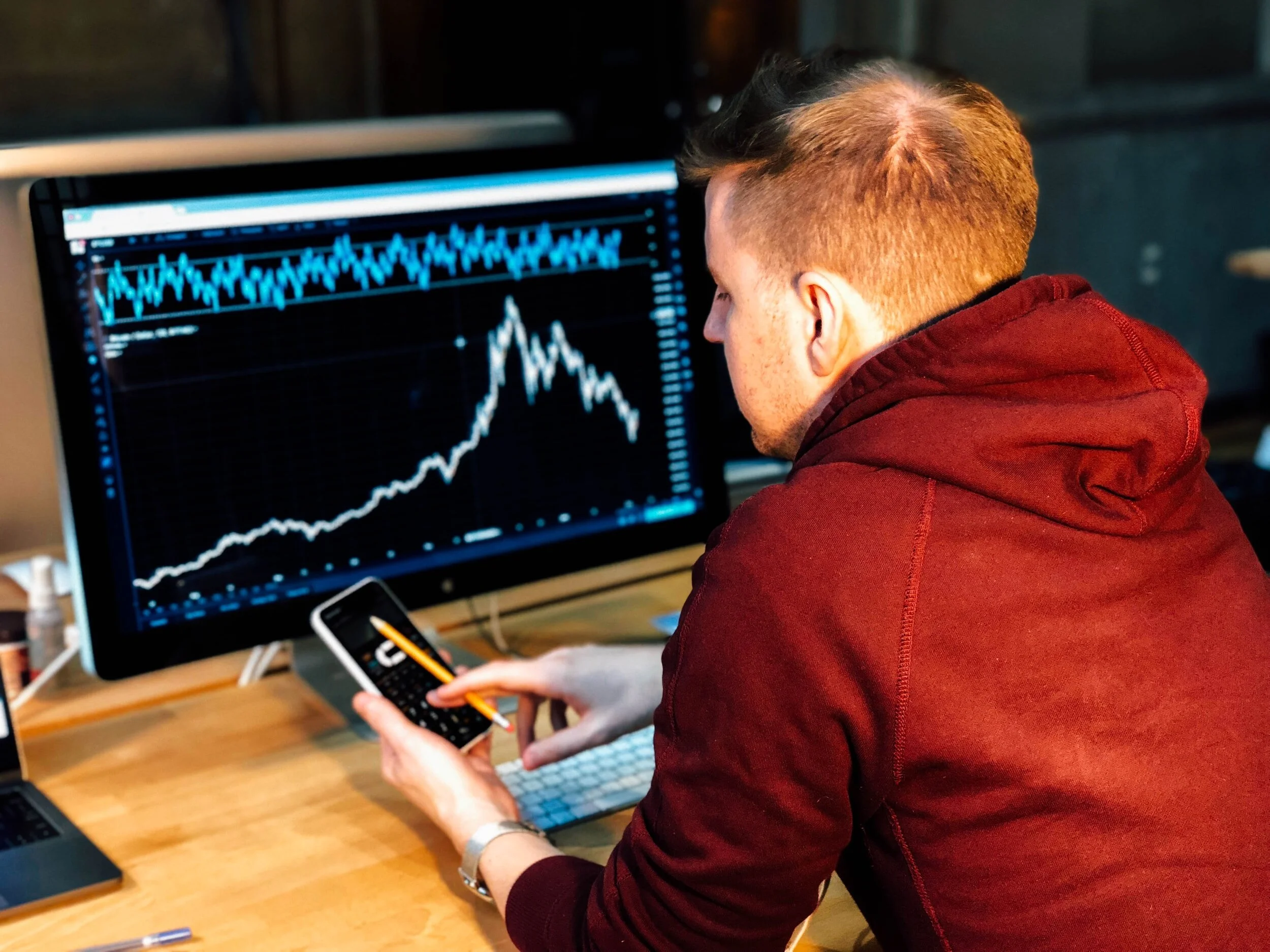 Person in a red hoodie analyzing a stock market graph on a computer monitor, using a smartphone calculator at a wooden desk.