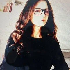 Person with long hair wearing glasses and a dark outfit indoors