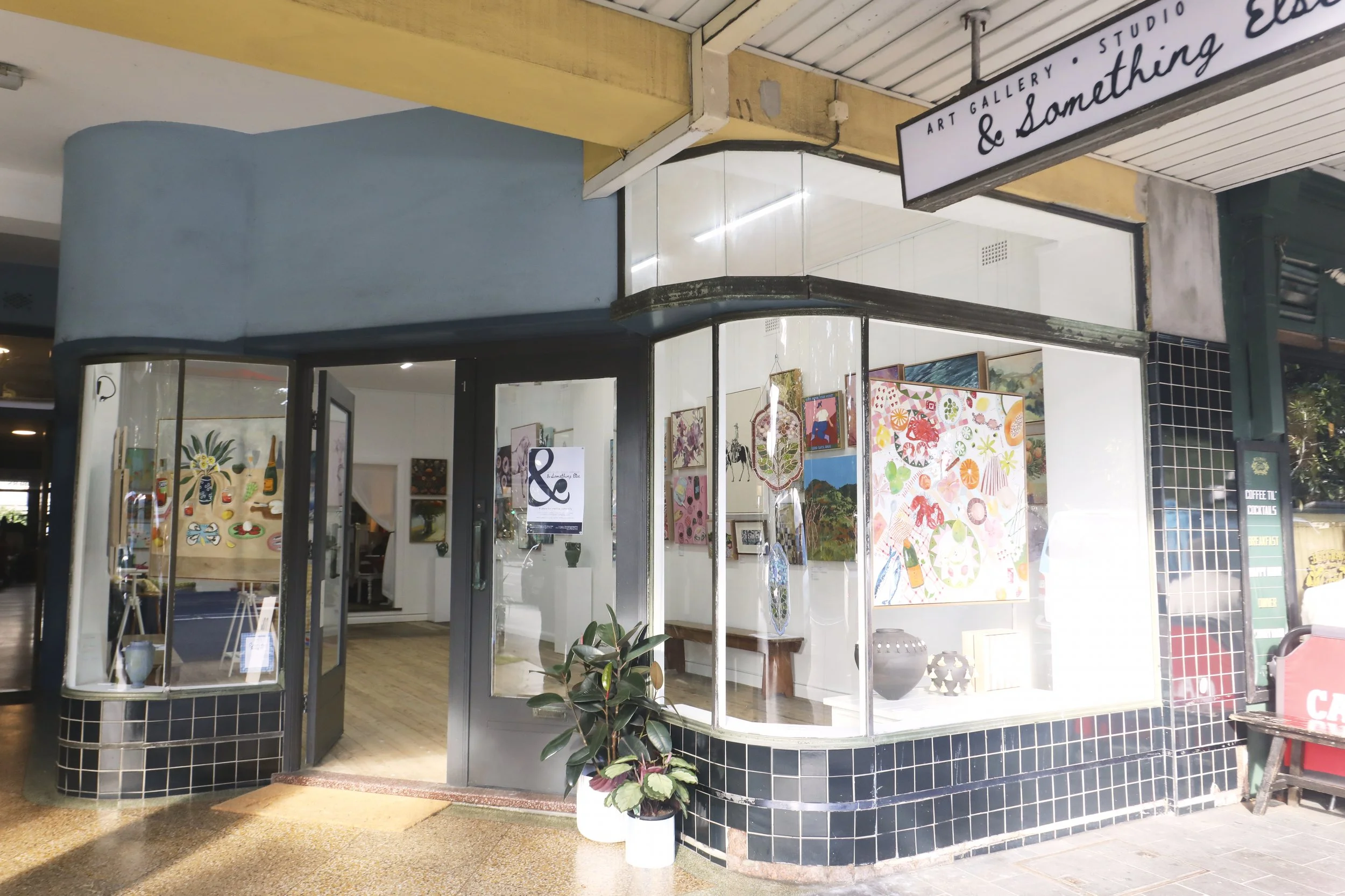 The storefront of an art gallery with large glass windows displaying colorful artwork inside. The gallery has a black tiled lower exterior and a pastel blue upper exterior with a curved corner entrance. A sign above reads 'Art Gallery & Something'.