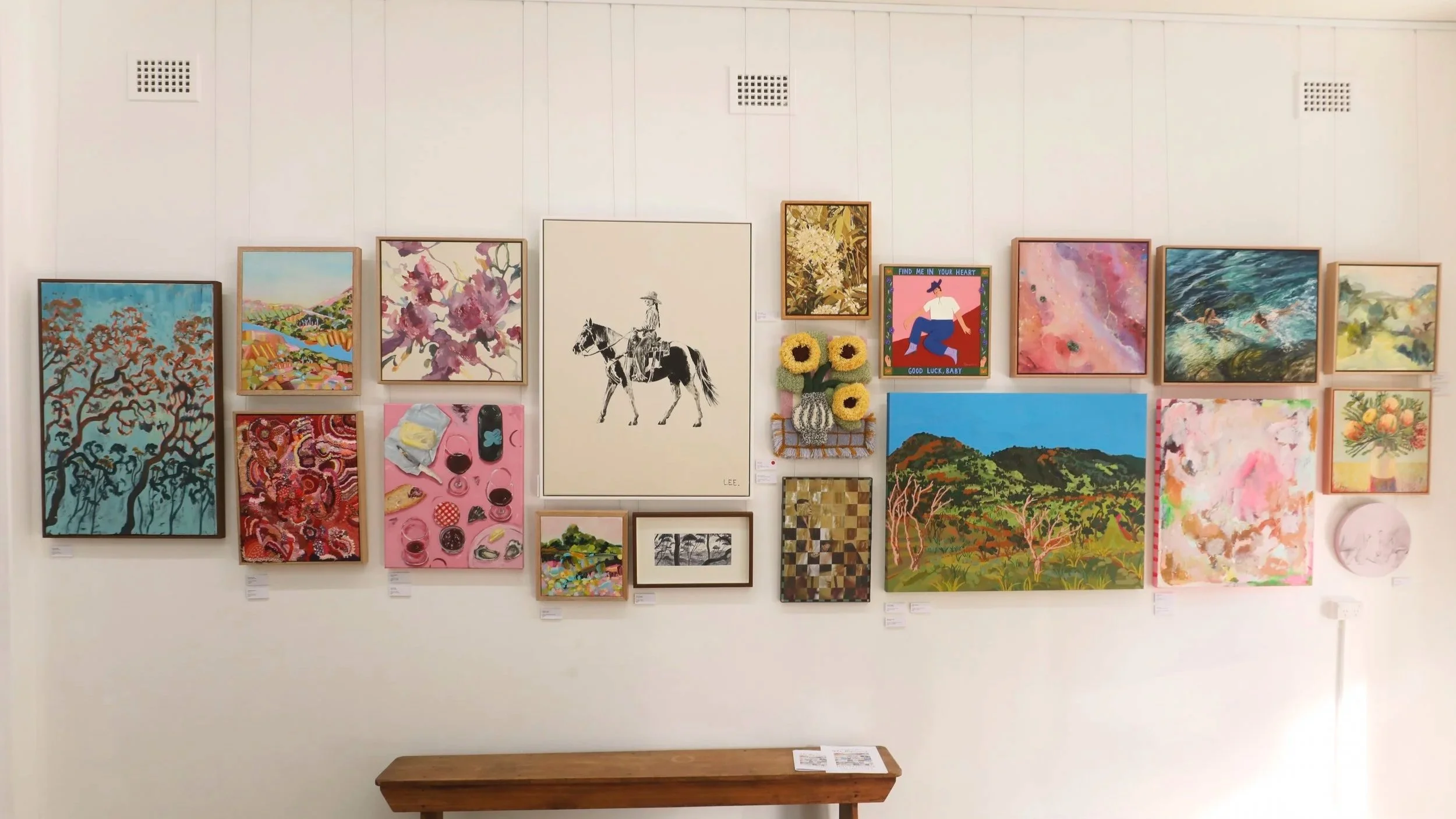 Gallery wall with multiple colorful paintings and artworks, including landscapes, abstract art, and a black and white picture of a person on horseback, with a wooden bench beneath.