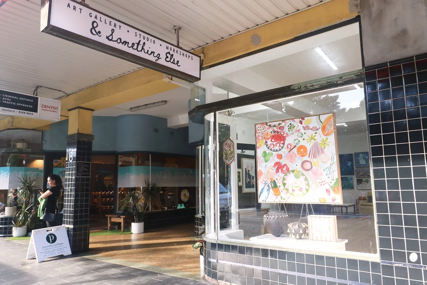 Exterior of an art gallery and studio with a sign that reads 'Art Gallery, Studio, Workshops & Something Else'. The gallery has a large window display with colorful artwork and interior furnishings visible through the glass. A woman wearing a face mask stands outside near potted plants and a white sign with a logo and text.
