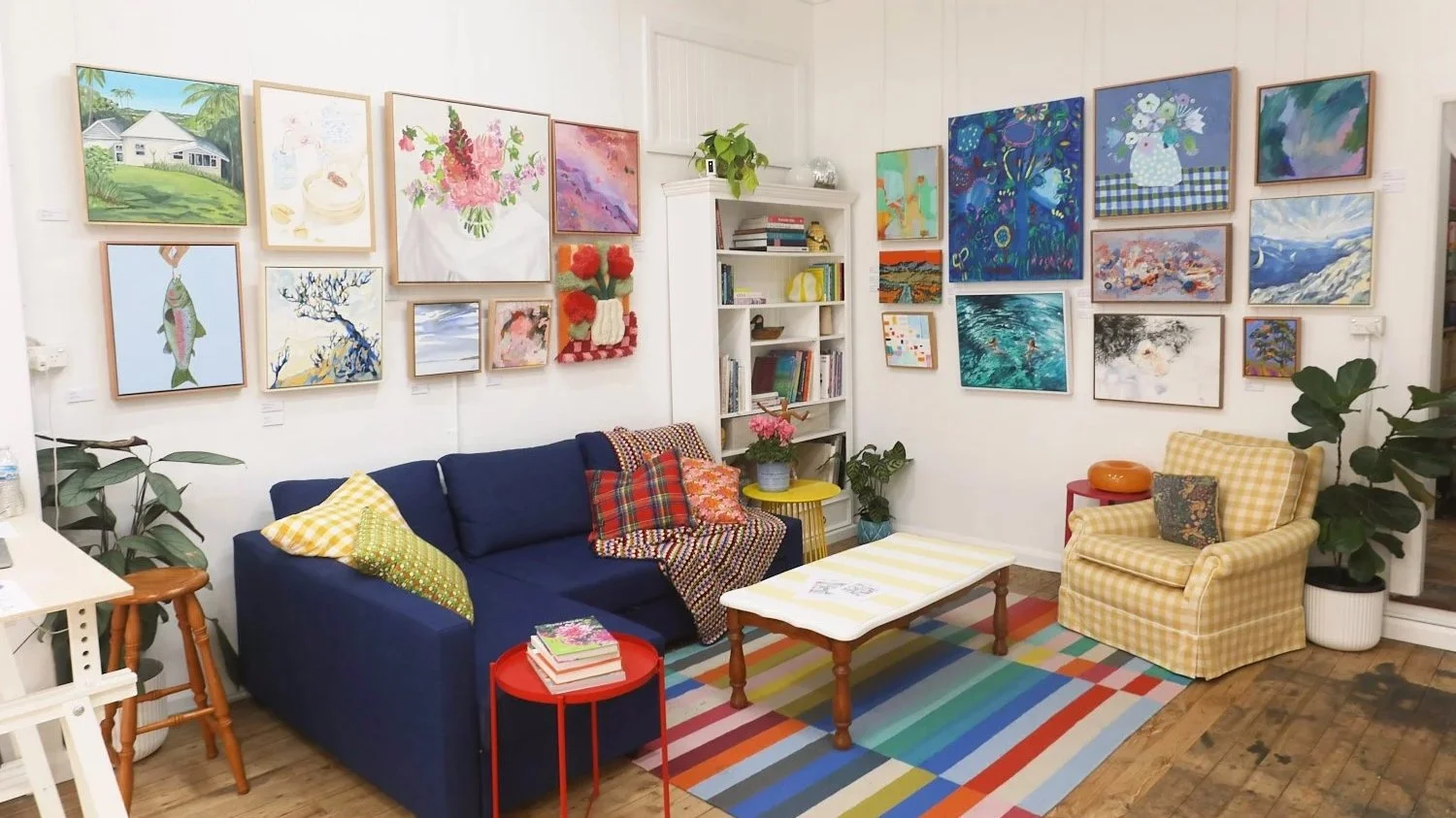 A cozy living room with a white wall decorated with numerous colorful paintings and illustrations. There's a navy blue sofa with patterned and solid throw pillows, a yellow-checkered armchair with a dark pillow, and a multicolored striped rug on a wooden floor. A small white coffee table, a red side table with books, a yellow side table, and several houseplants complete the scene. A white bookshelf filled with books and decorative items stands against the wall.