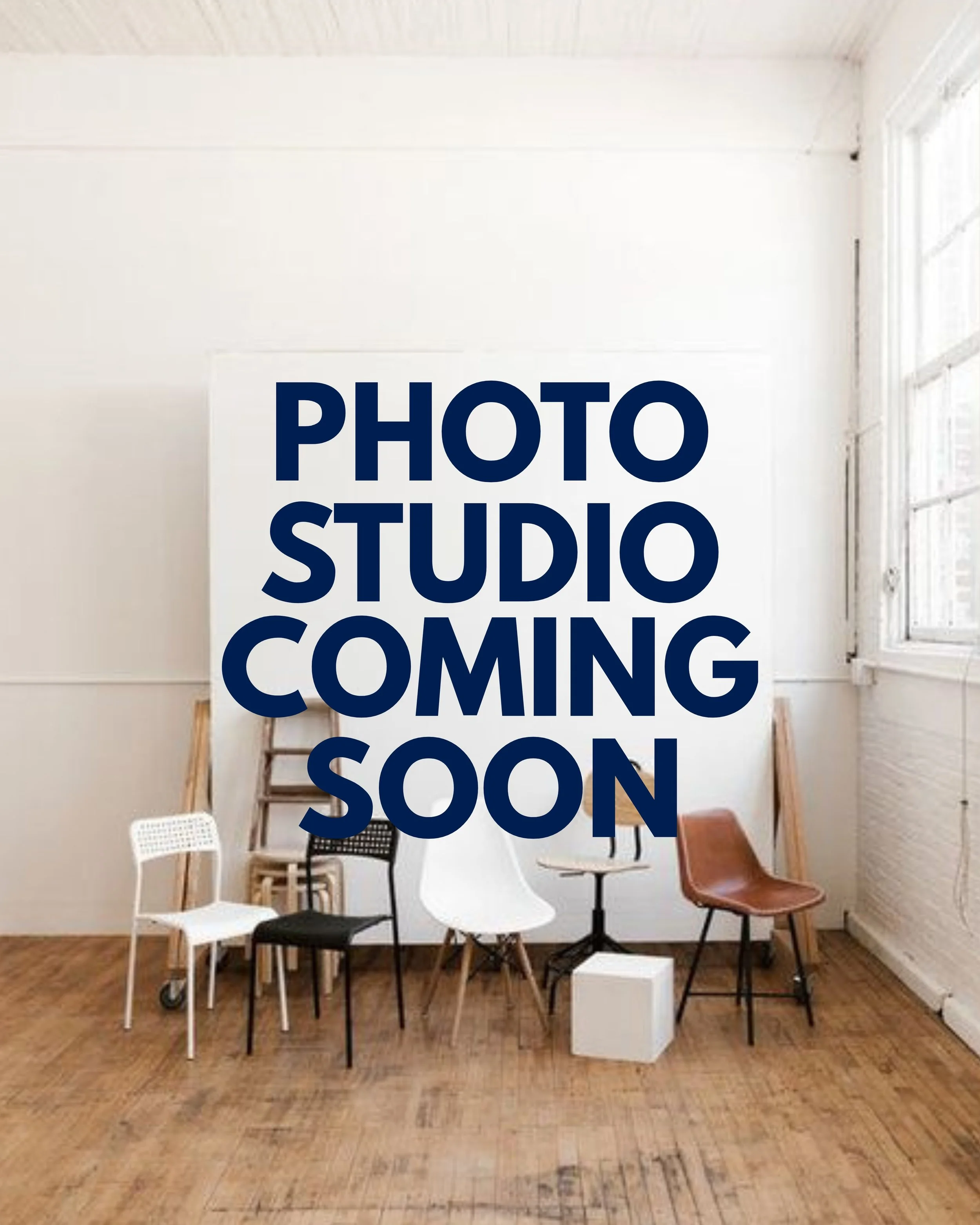 Empty photo studio with a white wall, wooden floor, and four chairs of different styles, with a large sign that reads 'Photo Studio Coming Soon' in bold blue letters.