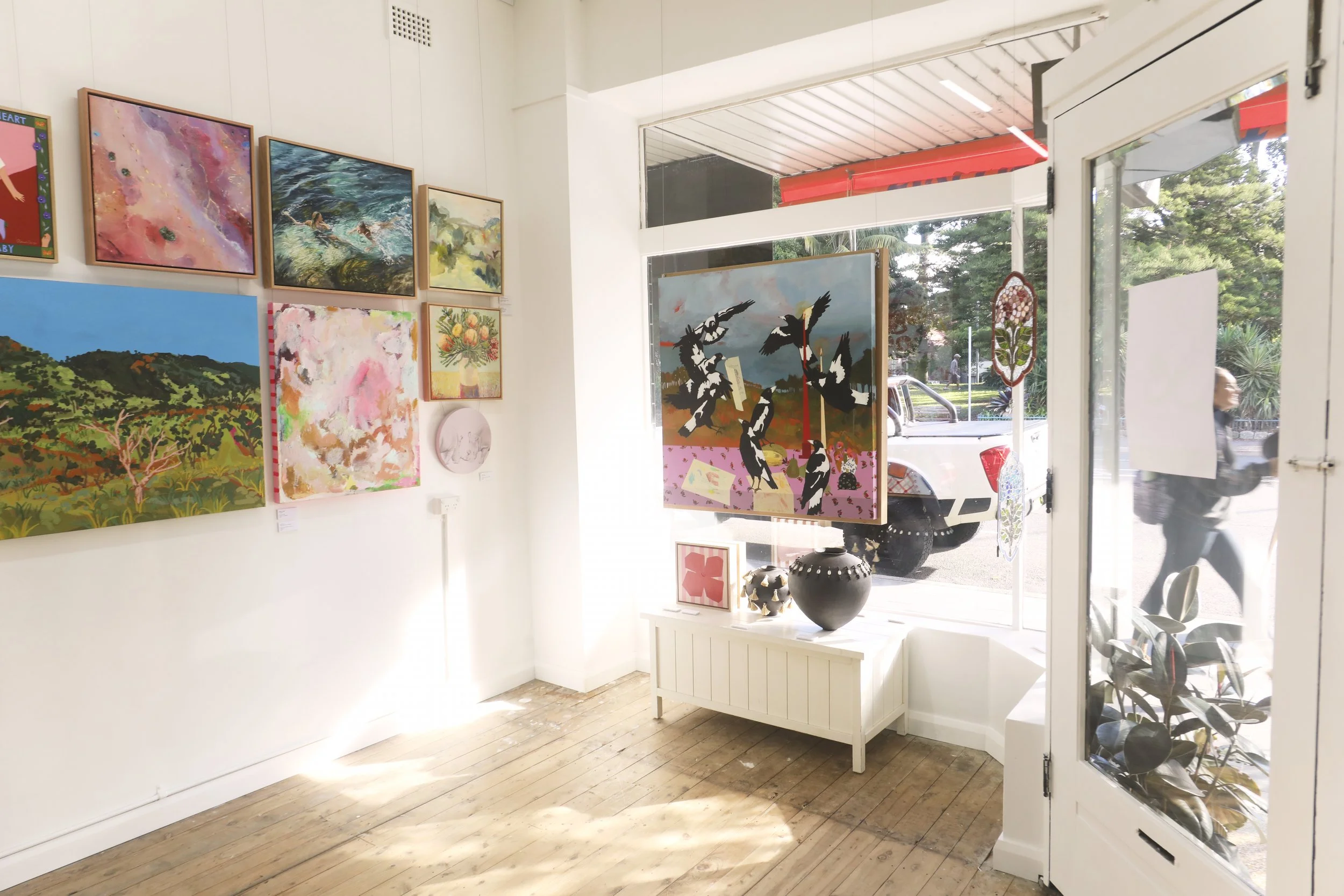 Interior view of an art gallery displaying multiple paintings on a white wall, with a view outside through the open glass door showing a street and greenery.
